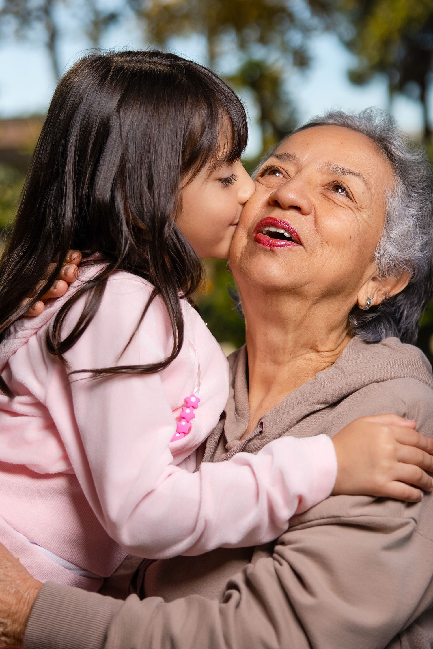 A young child in pink gives an affectionate kiss to an elderly person wearing a beige sweater outdoors.