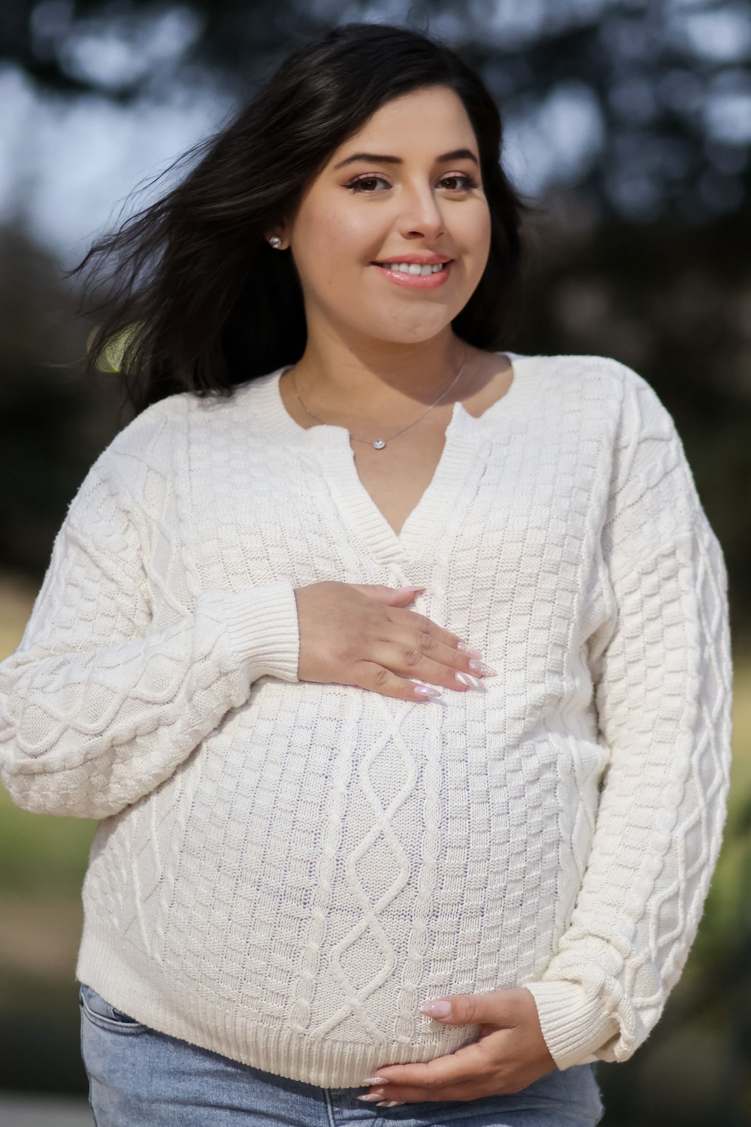 Woman in cozy white cable knit sweater poses outdoors with natural lighting and soft bokeh background.