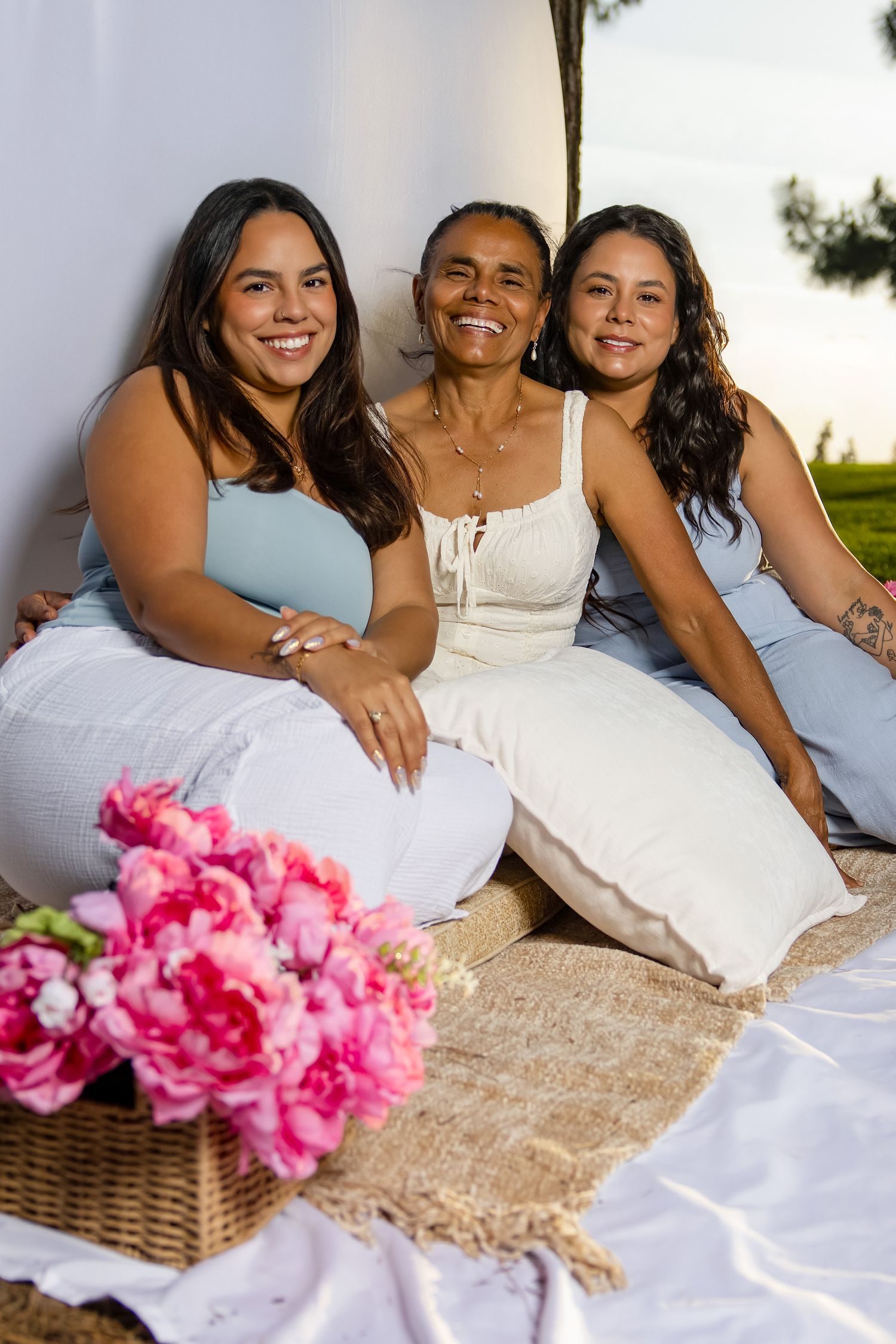 Three smiling women sitting together outdoors on a blanket with pink peonies in the foreground.