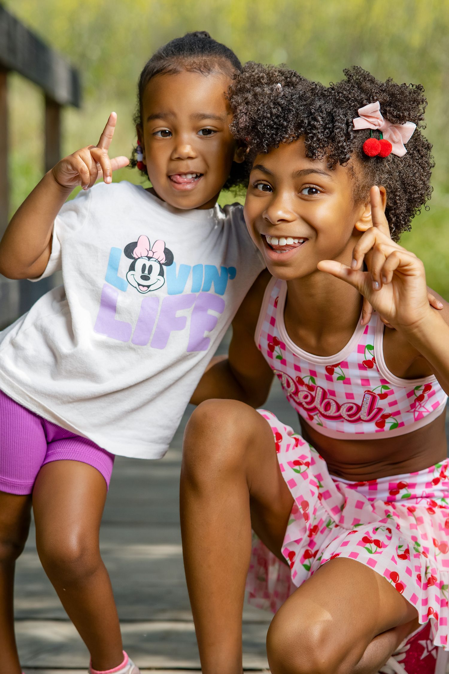 Two young girls pose outdoors on a wooden deck, one in a Minnie Mouse shirt and purple shorts, the other in a cherry print outfit.