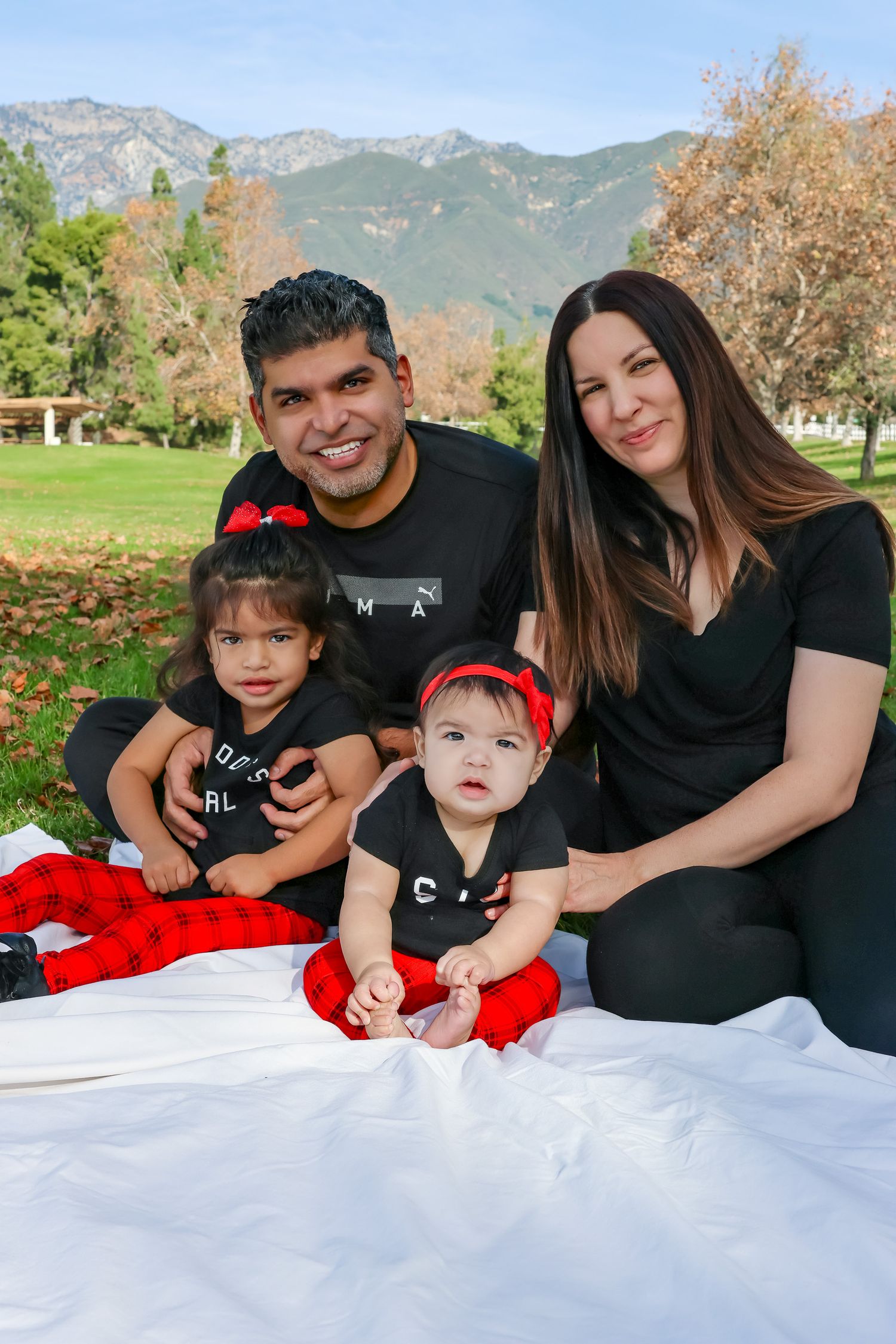 A family sits together on a white blanket for an outdoor photo with mountains and autumn trees in the background.