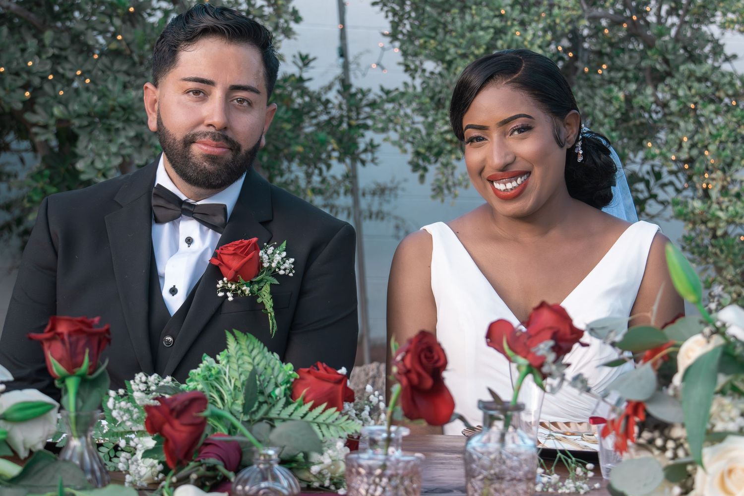 A wedding party poses together in front of greenery with red roses and white attire creating an elegant outdoor scene.