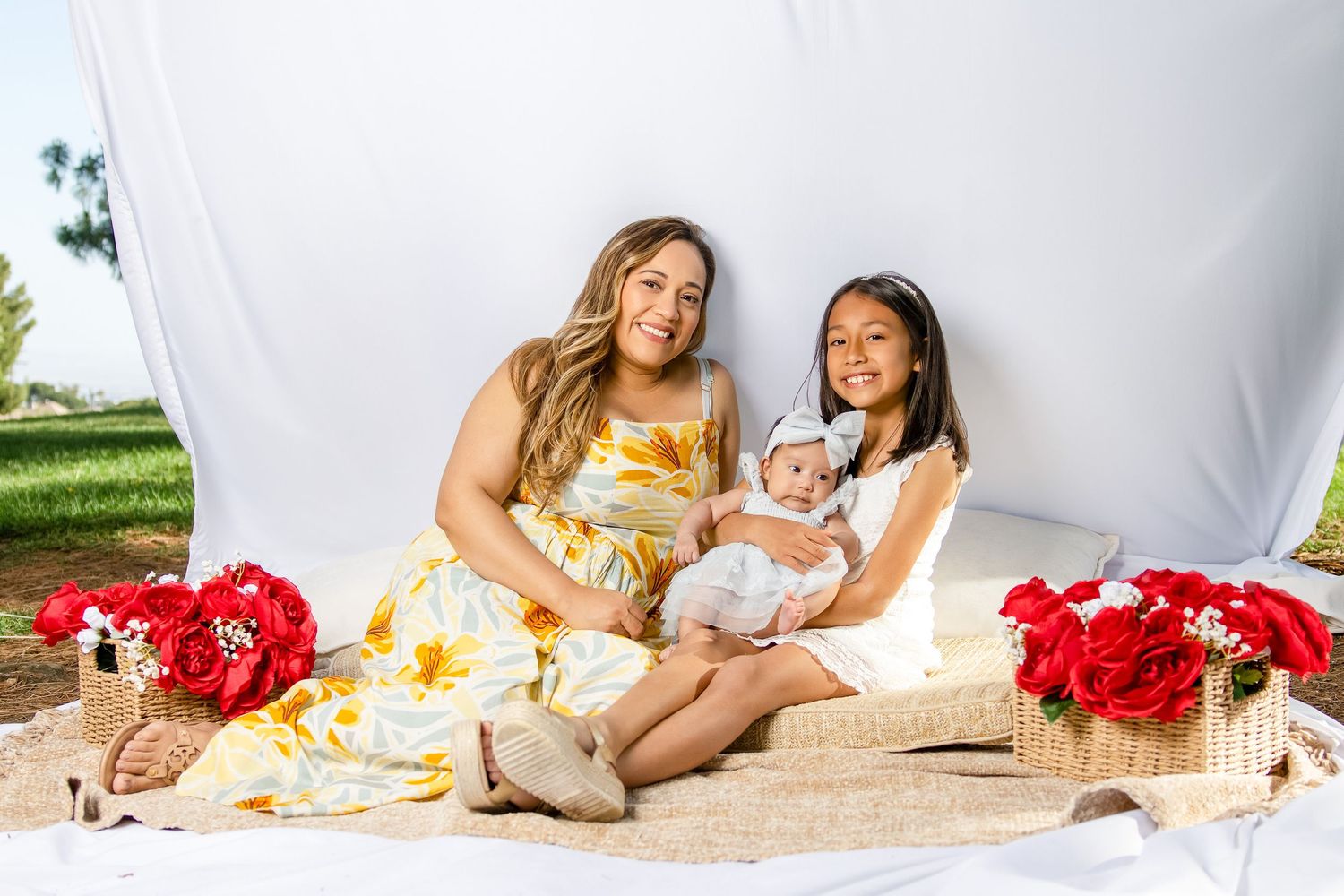 A woman in a floral dress sits with two girls, one holding a newborn, surrounded by red flowers outdoors.