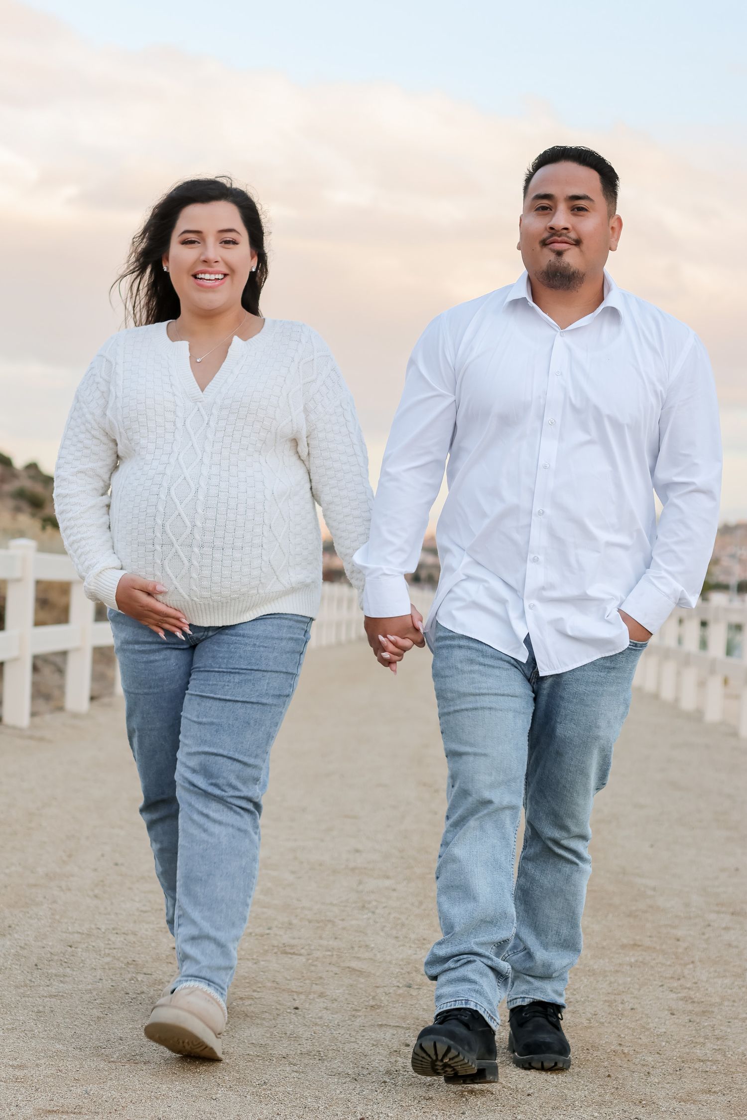 A couple holding hands walks along a sandy path by a white fence during sunset, wearing casual jeans and sweaters.