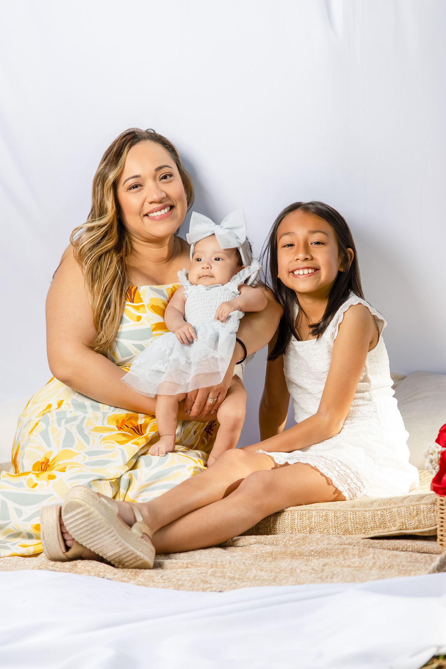 A smiling woman in a floral dress poses with a baby girl and young daughter on a white backdrop.