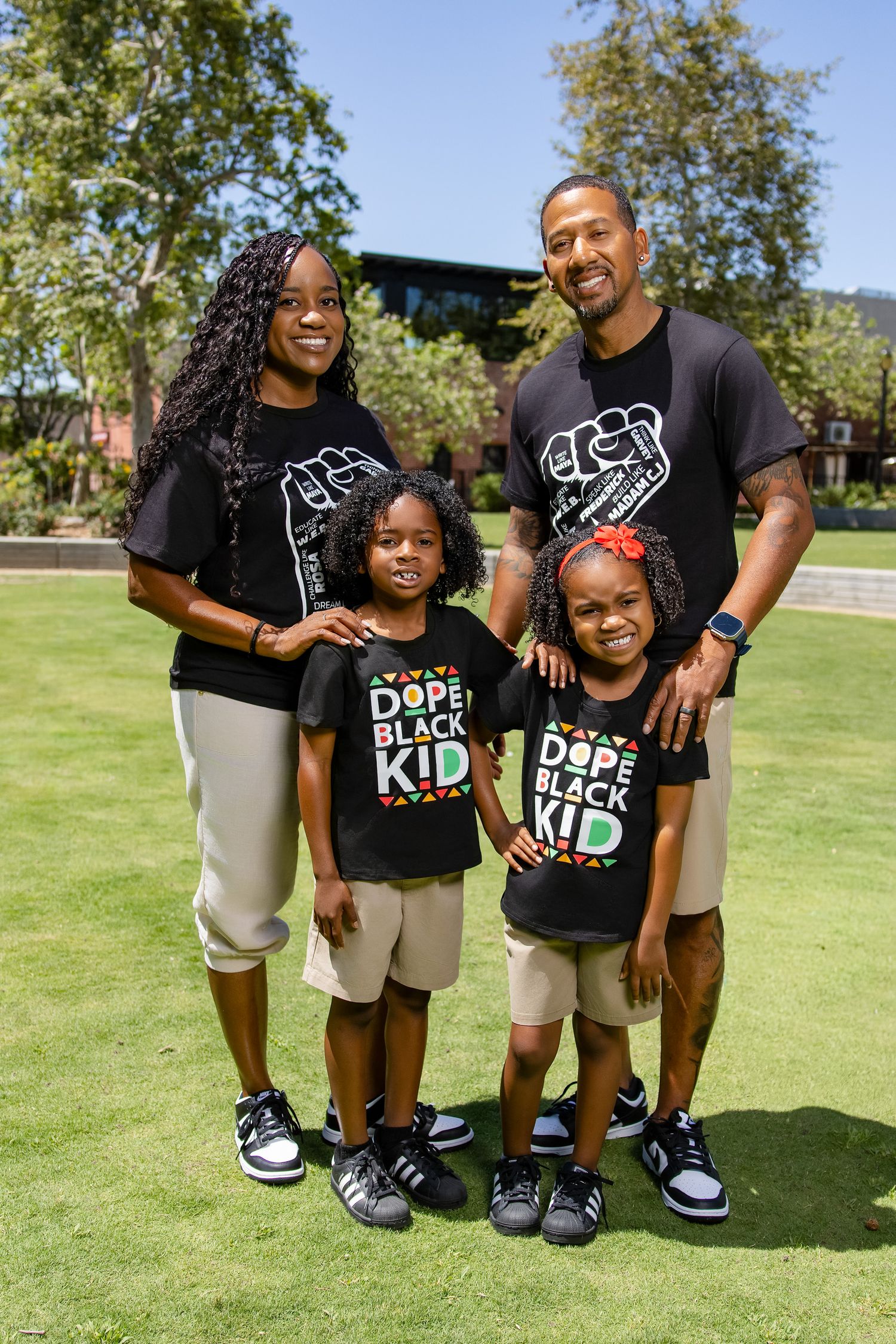 Family of four wearing matching black Dope Black Kid shirts smiling outdoors on green grass on a sunny day.