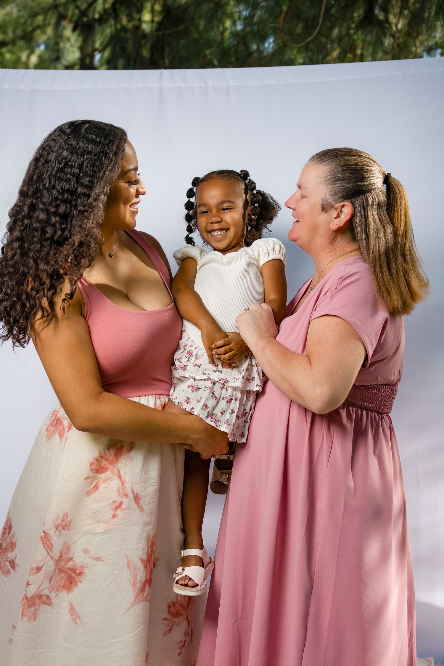 Two women in pink dresses joyfully hold a laughing little girl in a white floral outfit outdoors.