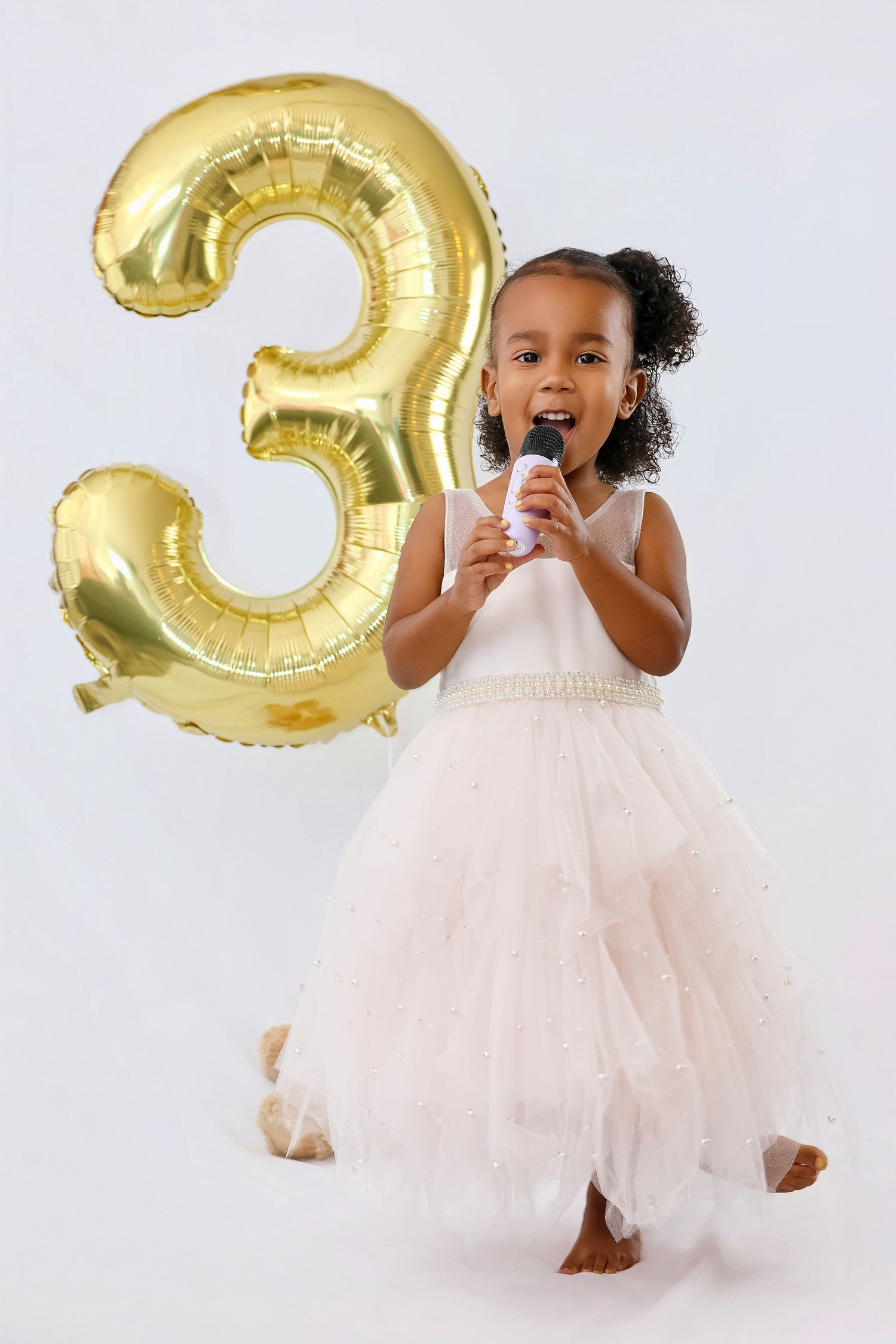 A toddler in a pink tulle dress poses with gold number balloons against a white backdrop.