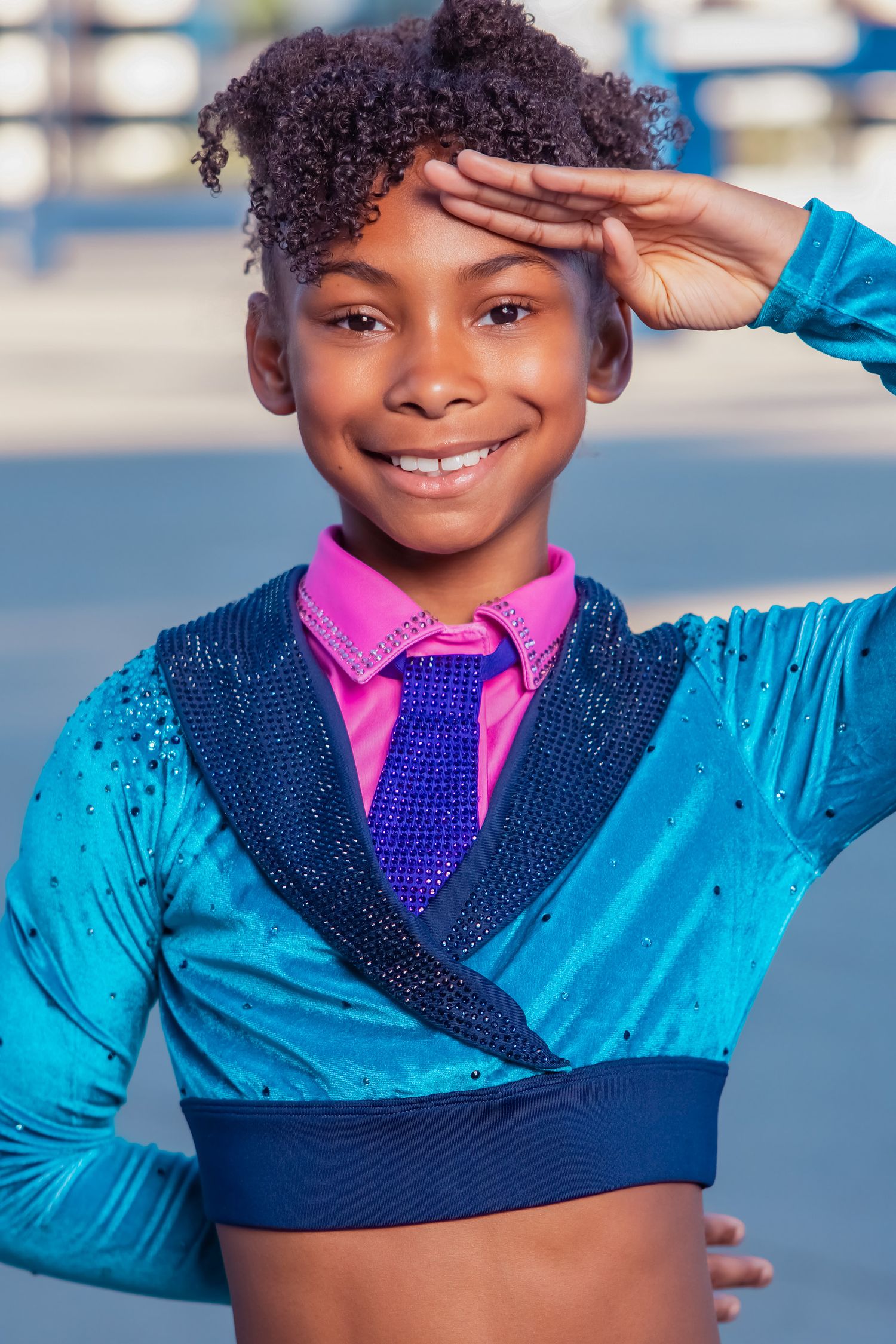Young girl in teal velvet dance costume with pink shirt and blue tie, smiling and saluting outdoors.