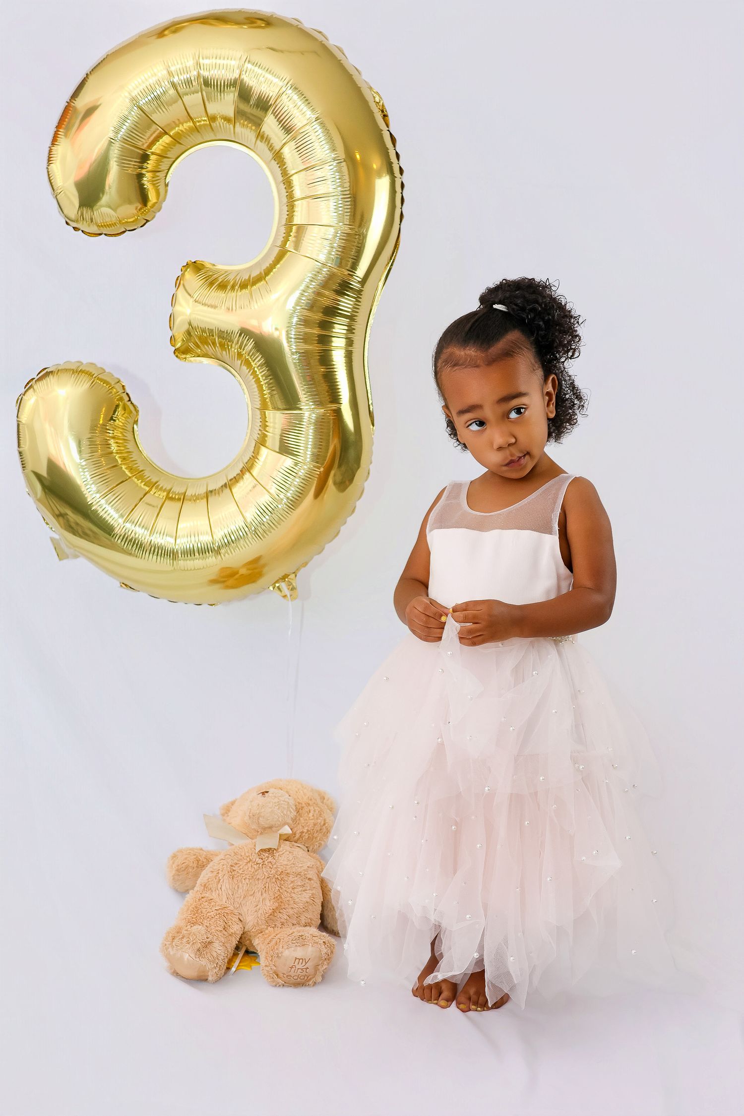 A child in a white tulle dress stands next to a large gold number 3 balloon and teddy bear against a white background.