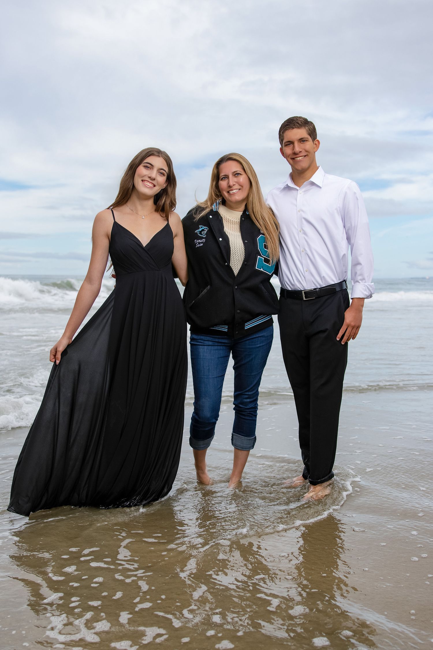 Three people smiling together on a beach, waves behind them, wearing formal and casual attire.