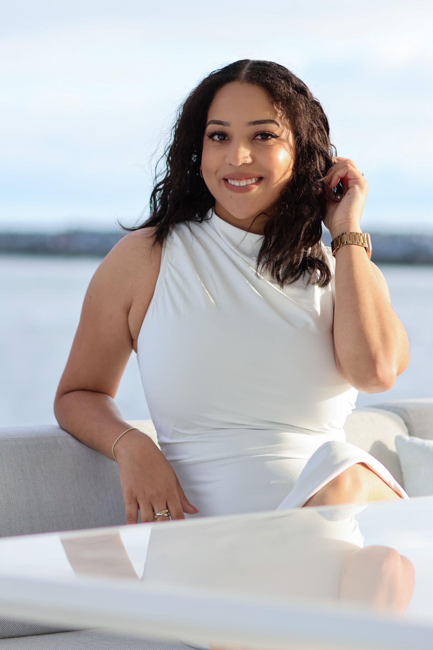 A woman in a white dress poses elegantly against a bright waterfront background.