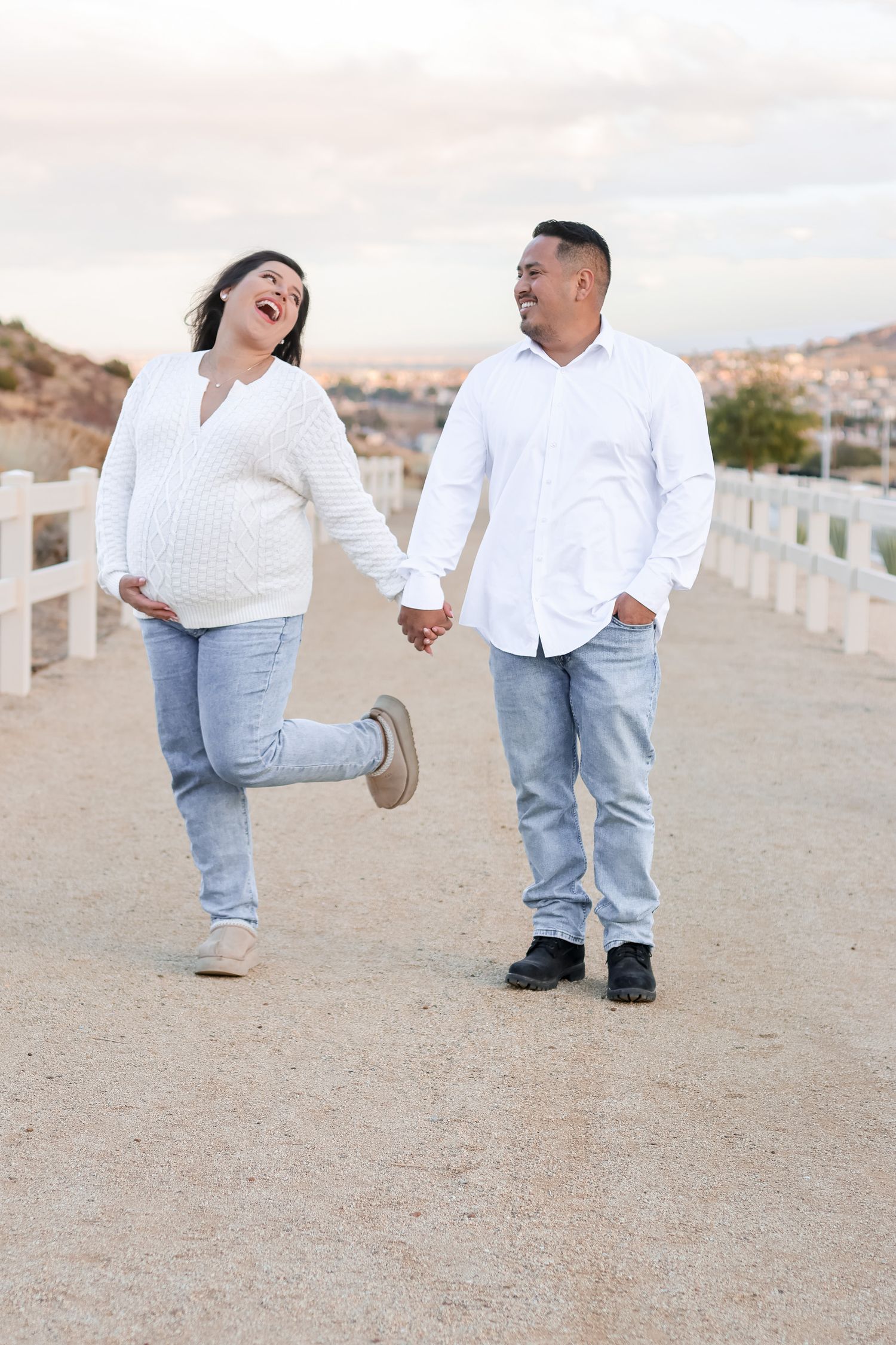 Couple in white shirts and jeans walking playfully along sandy path.