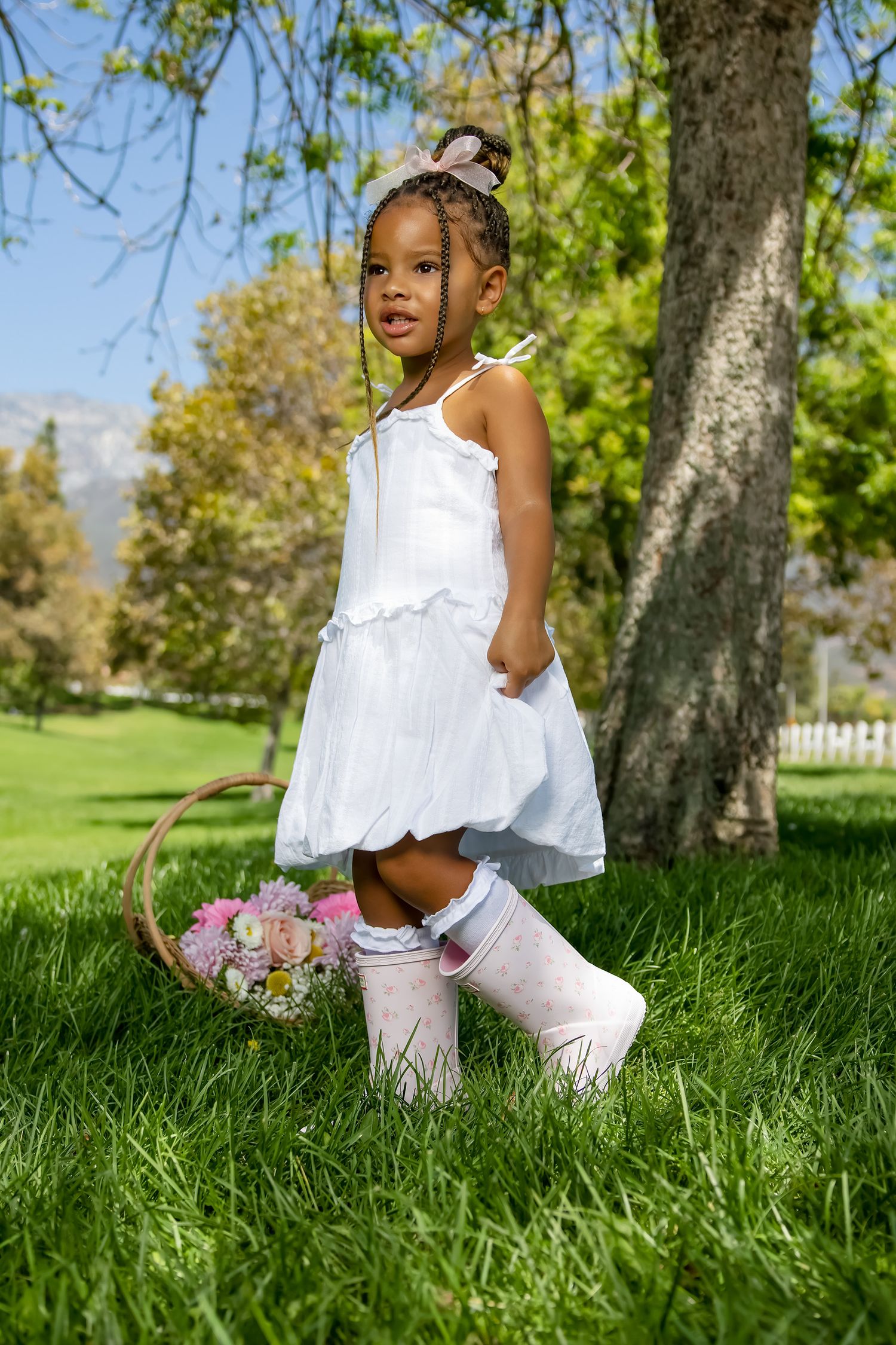Young Black girl in white dress and pink boots holds flower basket while standing on green grass near a tree outdoors.