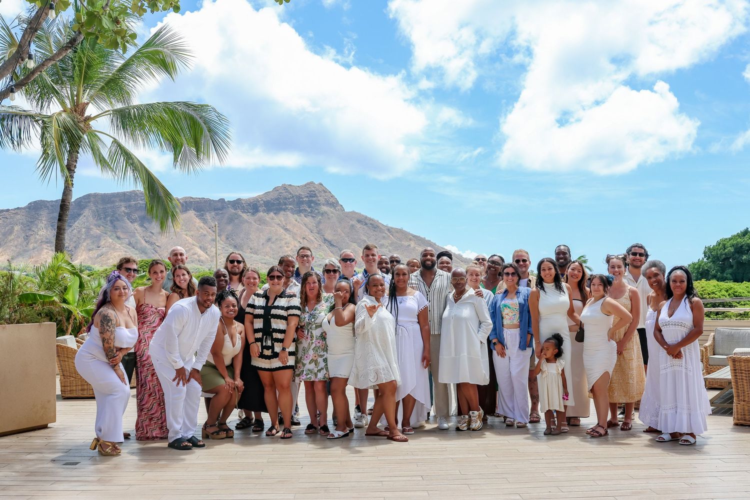 Panoramic beach group photo with people dressed in white against mountain and palm tree backdrop.