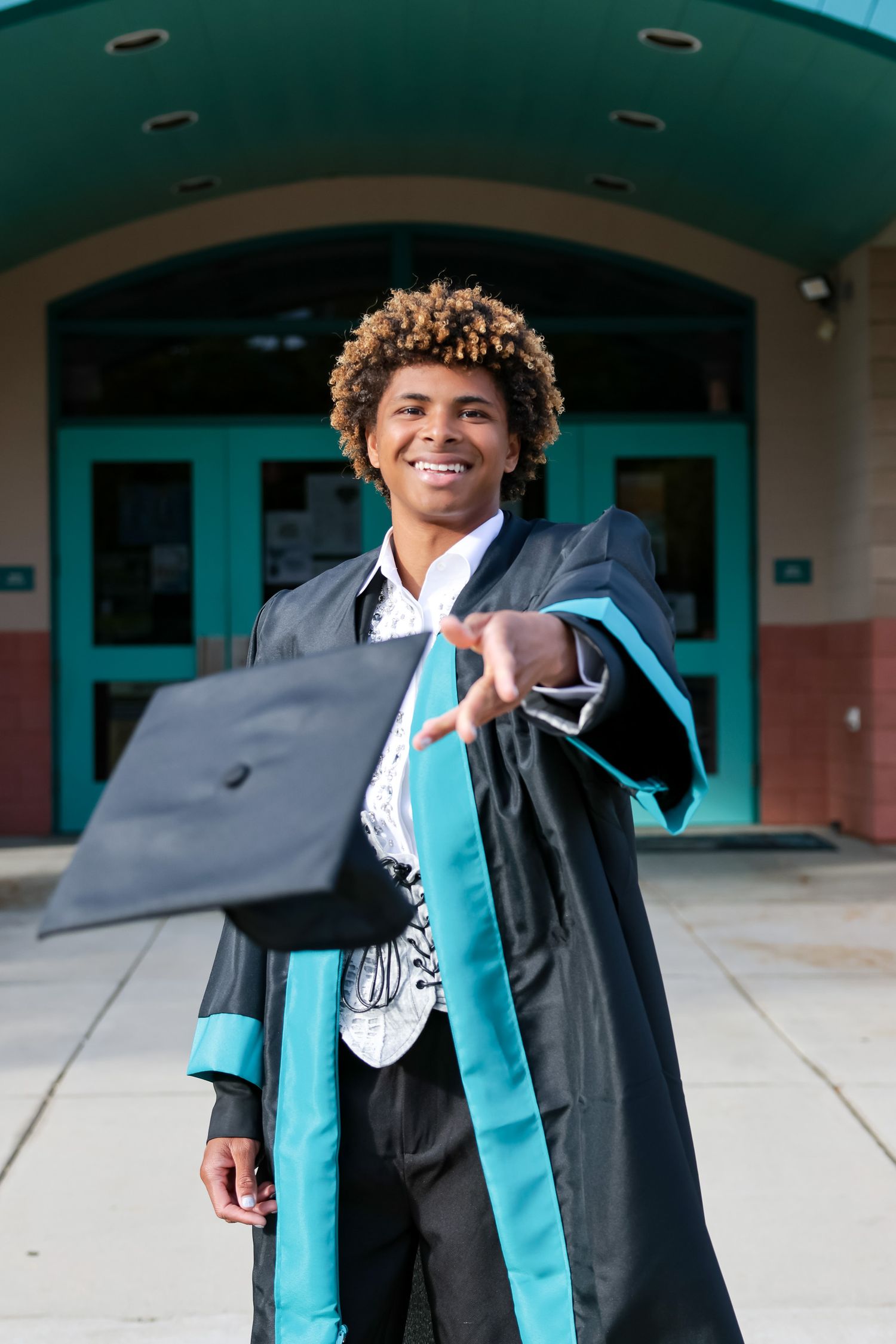 Graduate in black gown and teal stole poses joyfully outside school building.