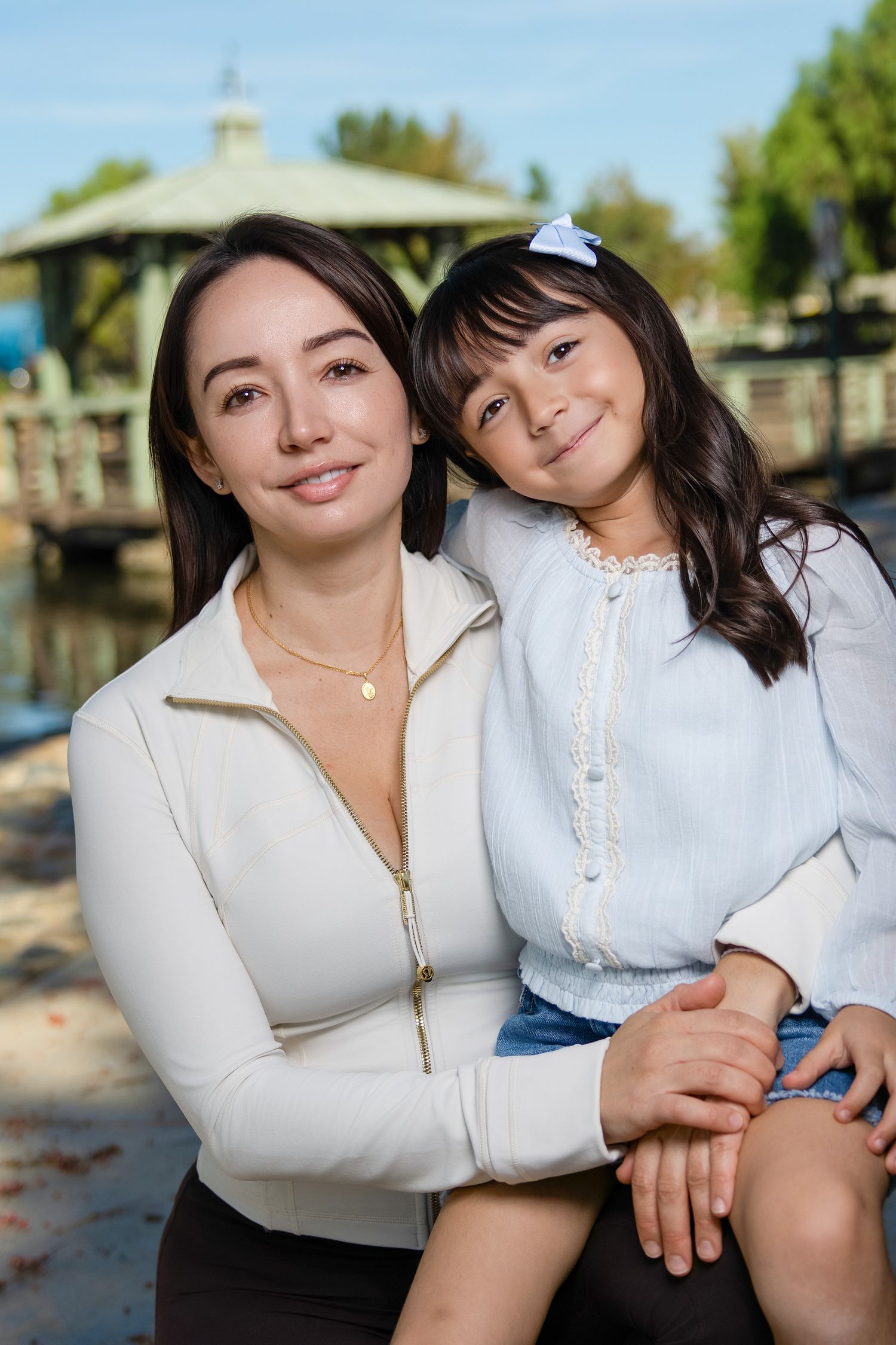 Mother and daughter in white tops sitting together by waterfront on sunny day.