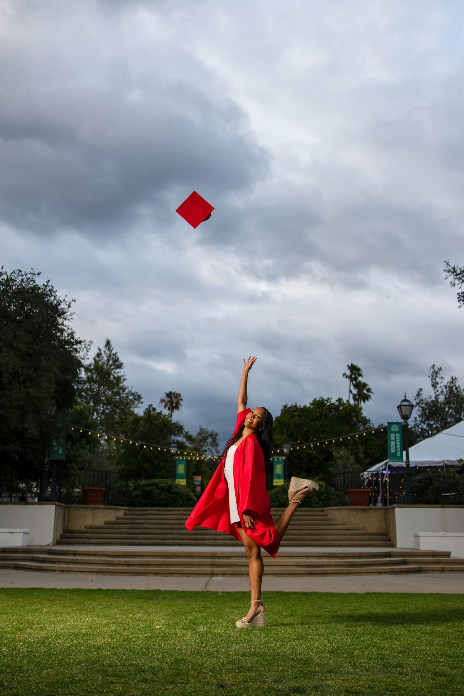 Graduate in red cap and gown joyfully tosses mortarboard into cloudy sky while jumping on green campus lawn.