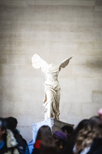 Ancient classical statue displayed in museum with crowd silhouettes below.