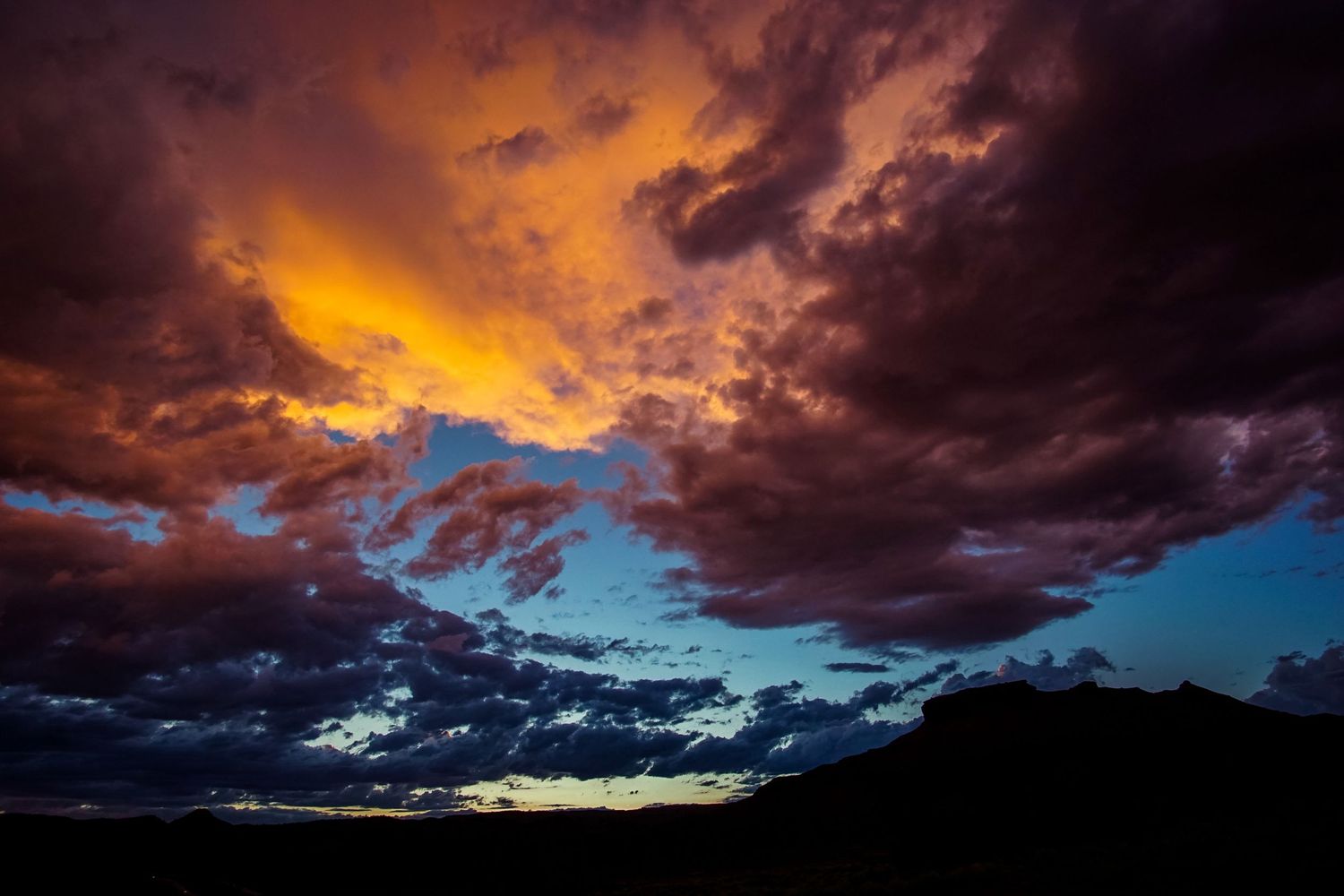 Dramatic sunset sky with orange and blue clouds silhouetting mountain landscape.