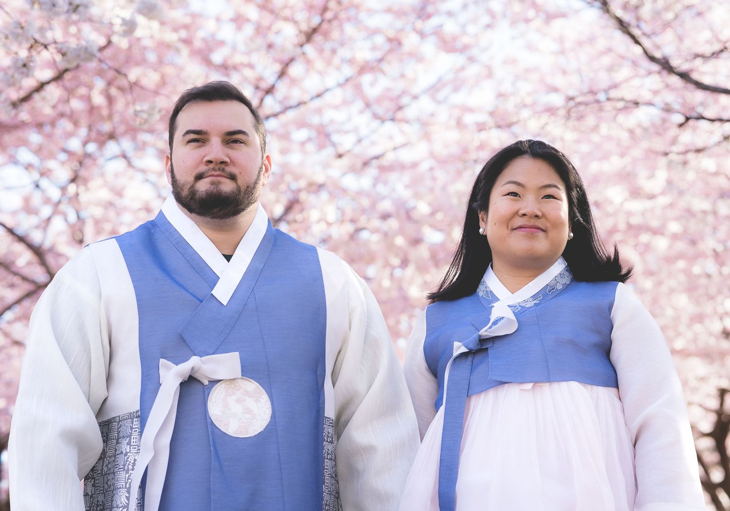 Couple wearing traditional Korean hanbok posing under cherry blossom trees.