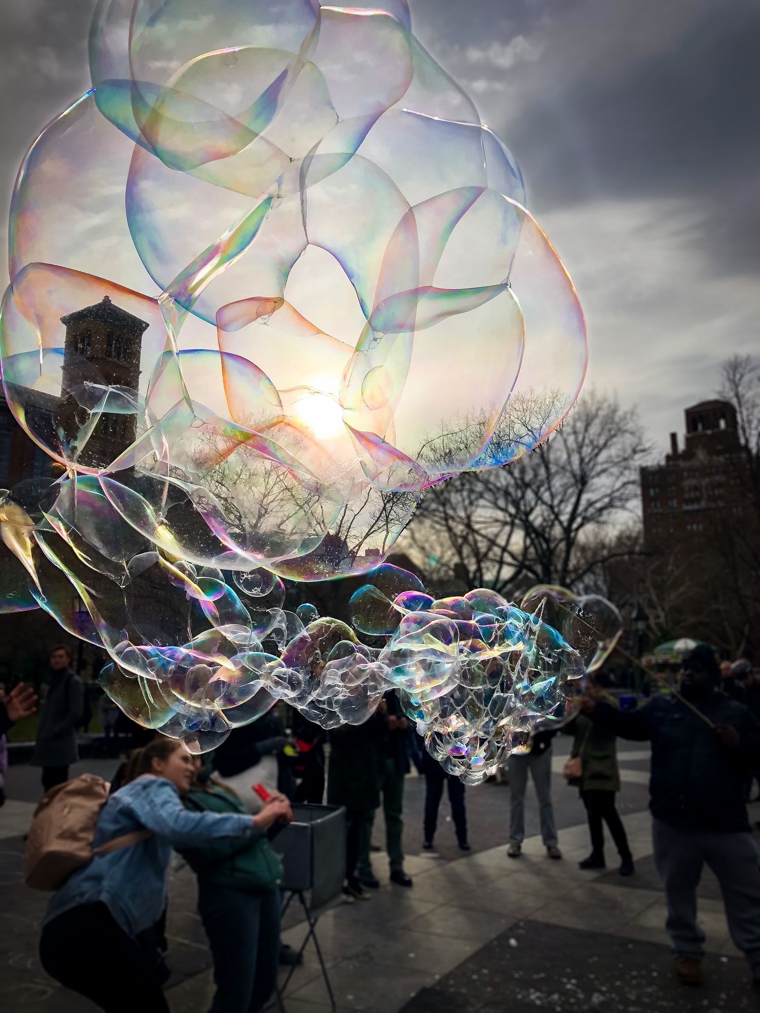 Large iridescent soap bubbles float against dramatic cloudy sky in city park.