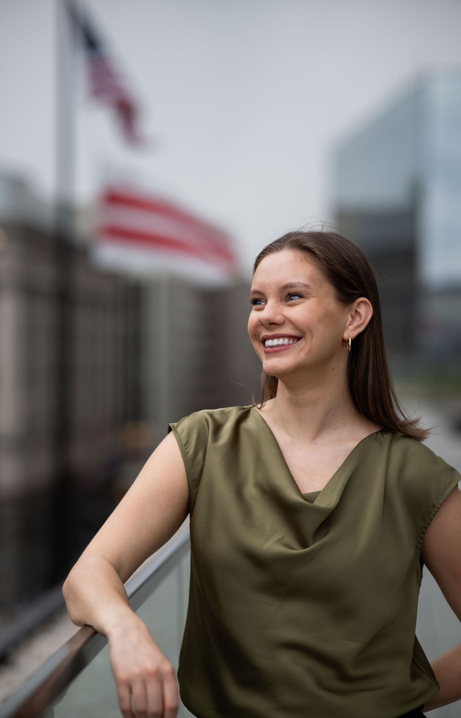Professional in olive sleeveless top poses cheerfully with cityscape and flags in background.