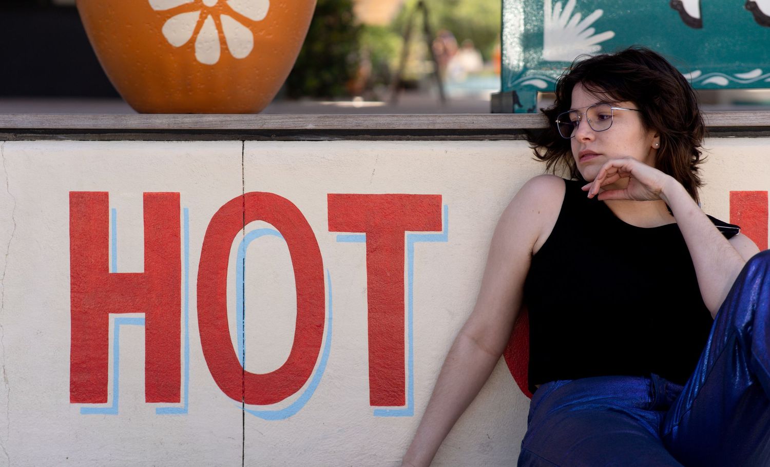 Young woman with glasses sitting beside a bold red HOT sign outdoors, with a pumpkin decoration in the background.