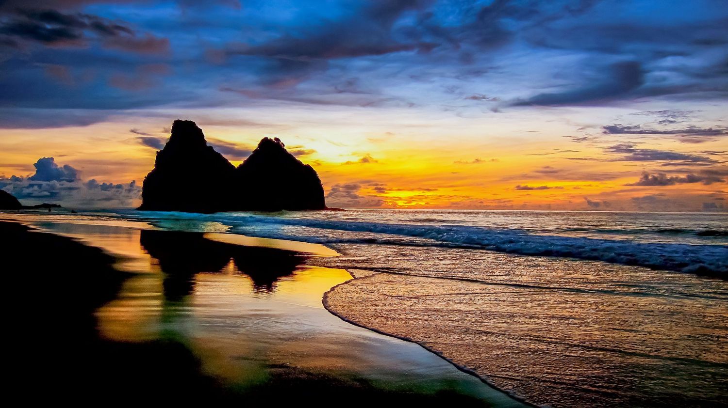 Dramatic coastal sunset panorama with silhouetted rock formations and colorful sky reflections on wet sand.