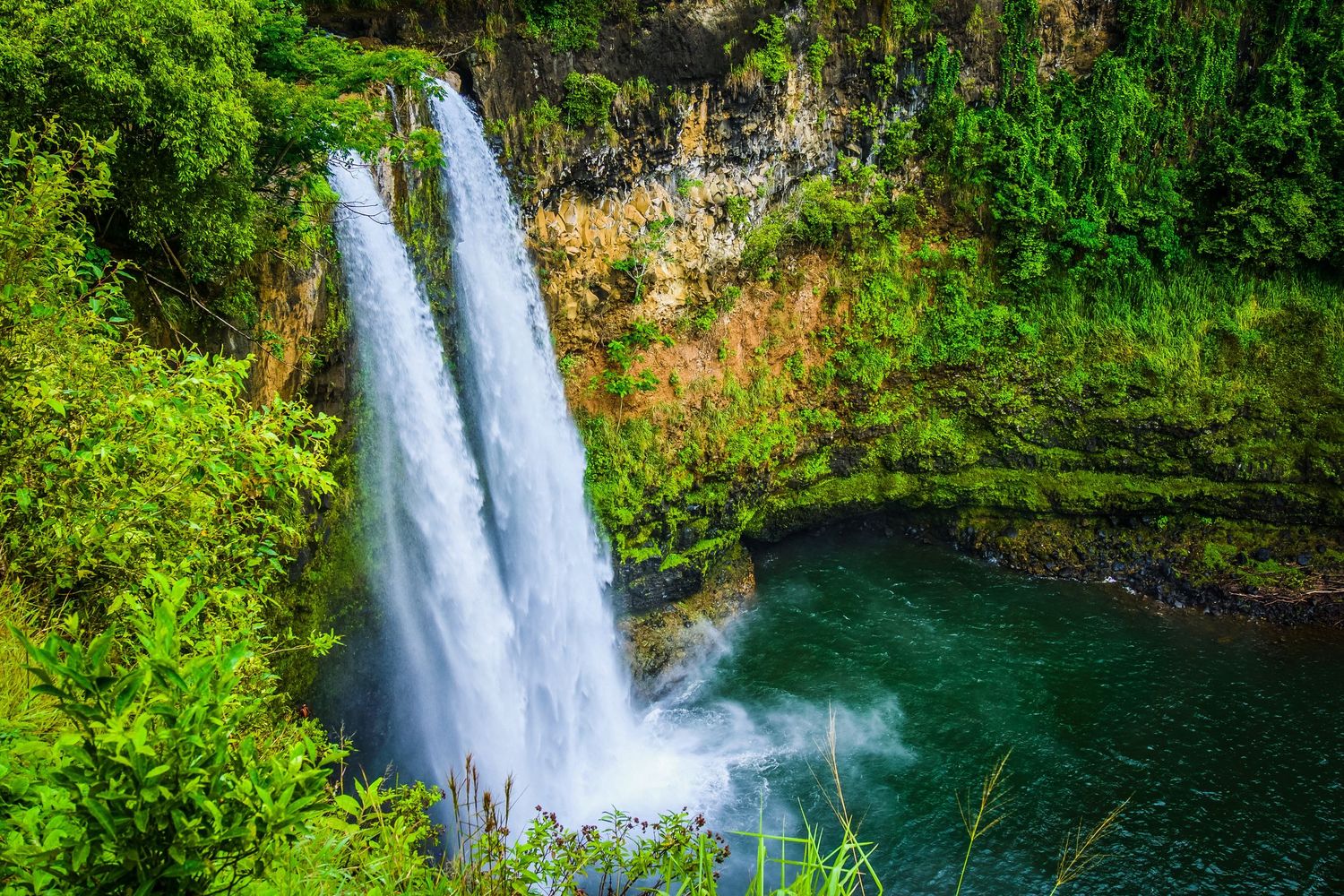 Majestic twin waterfalls plunge into emerald pool surrounded by lush Hawaiian vegetation.