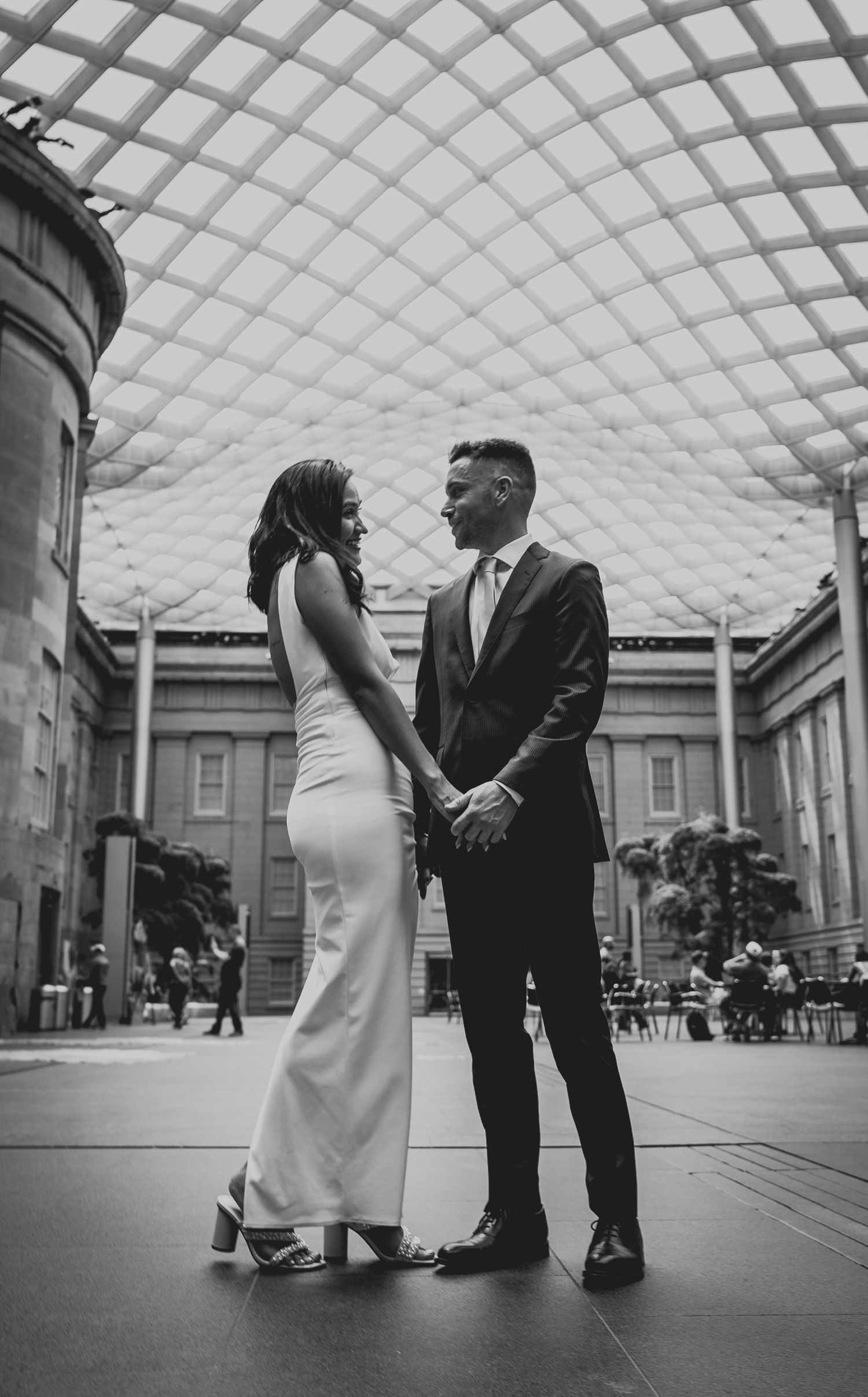 Couple in wedding attire holding hands in elegant domed atrium, black and white photo.