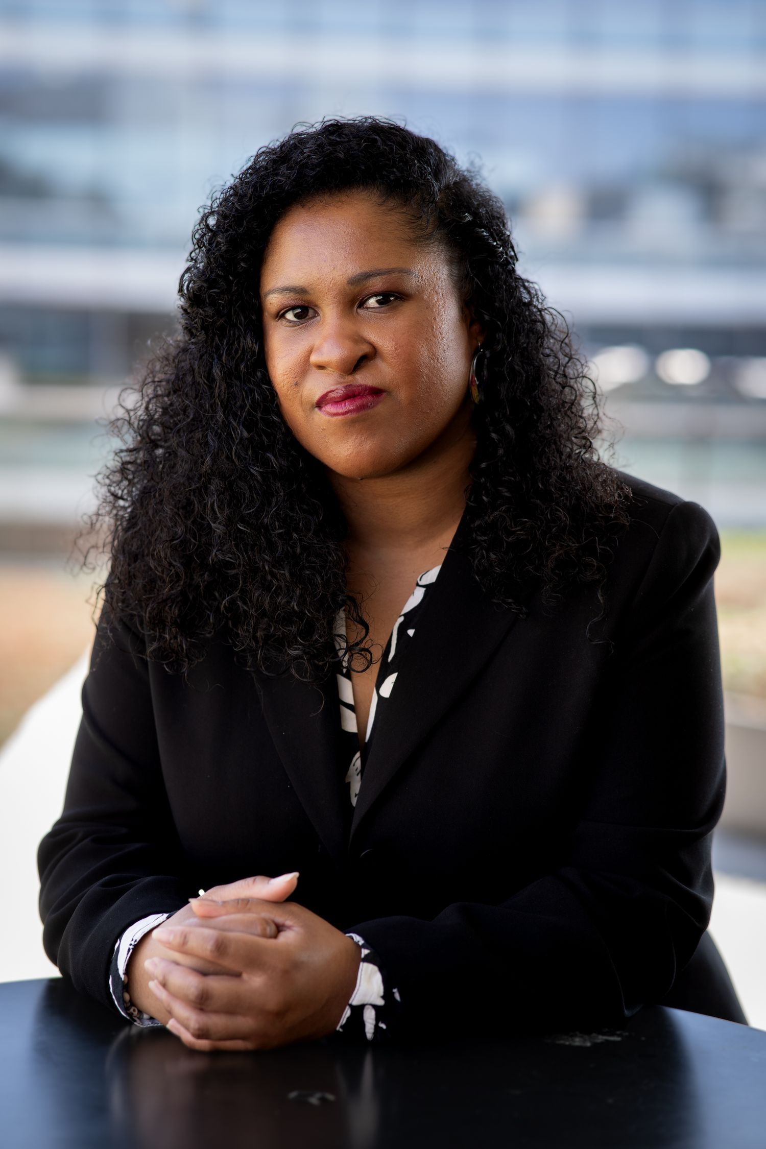 Professional Black woman with curly hair in black blazer sits at table with hands clasped in corporate setting.