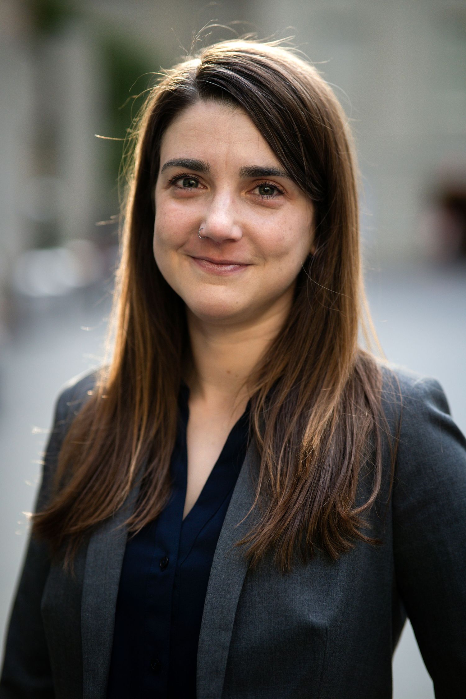 Professional with long brown hair wearing dark blazer smiles warmly in outdoor portrait.