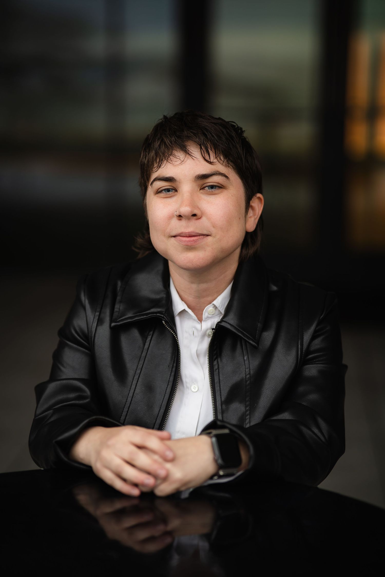 Individual wearing leather jacket poses casually in dimly lit environment.