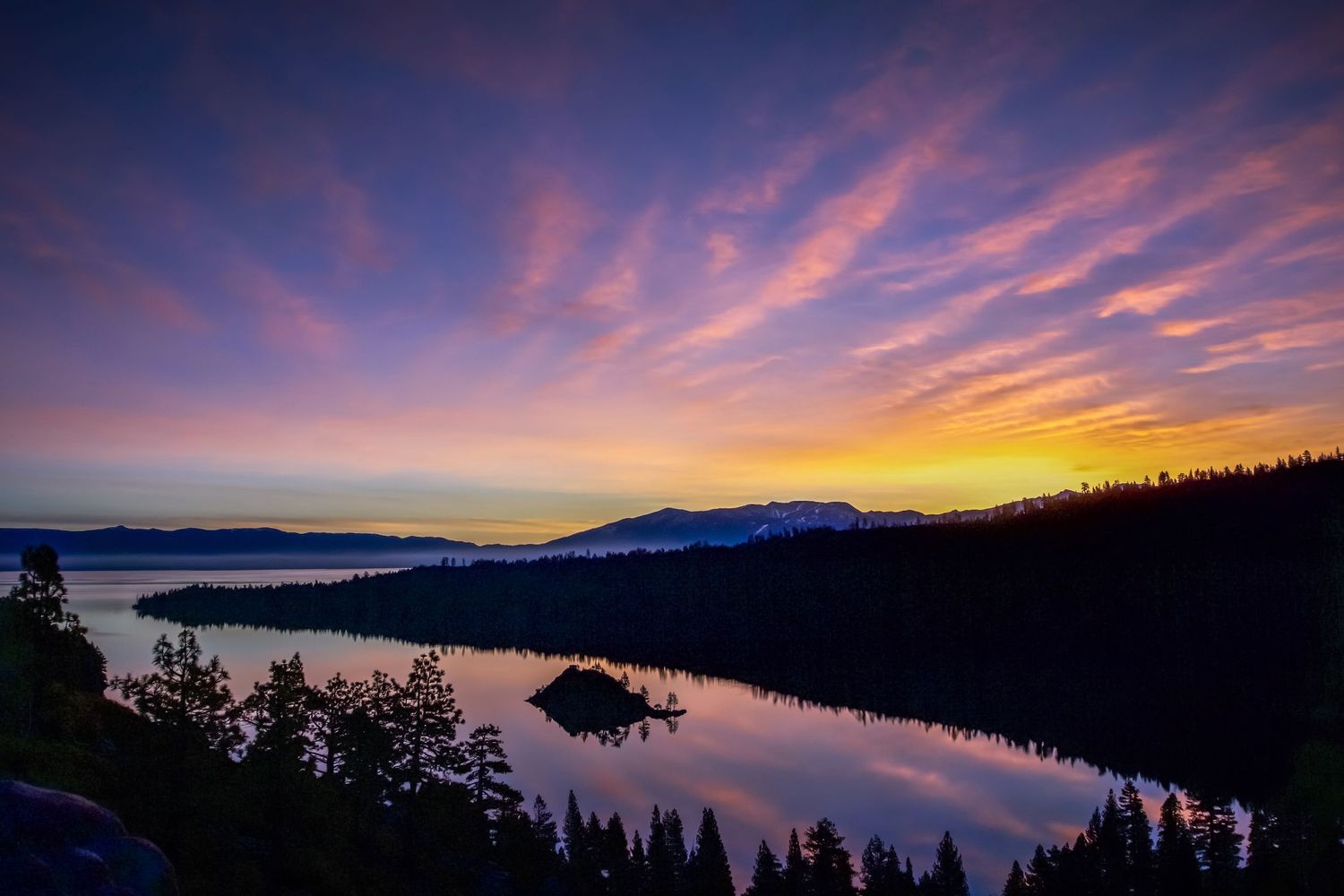 Stunning purple and orange sunset sky over a mountain lake with silhouetted pine trees.