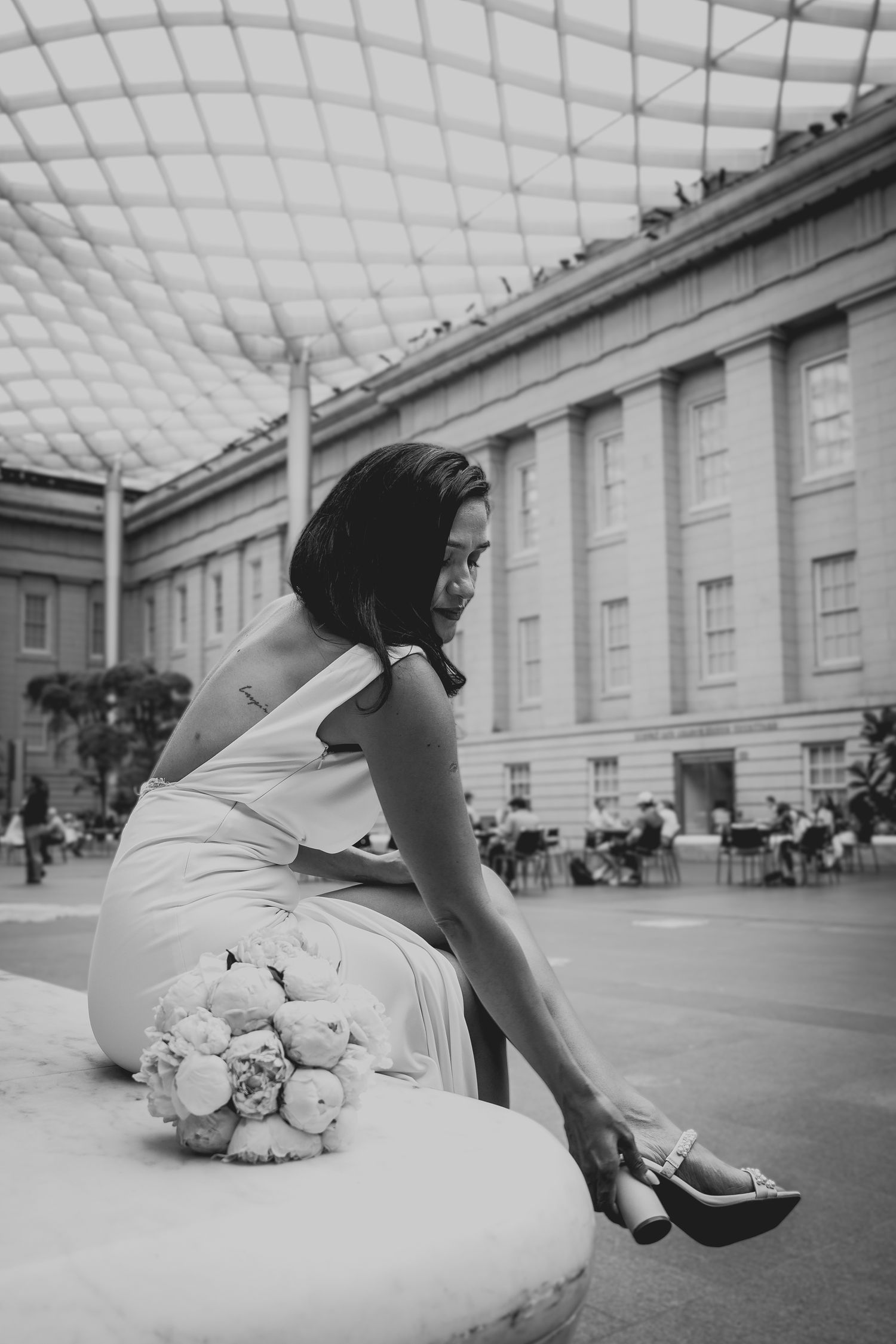 Black and white photo of a bride in a white dress adjusting her heel, with a bouquet beside her in an elegant venue.