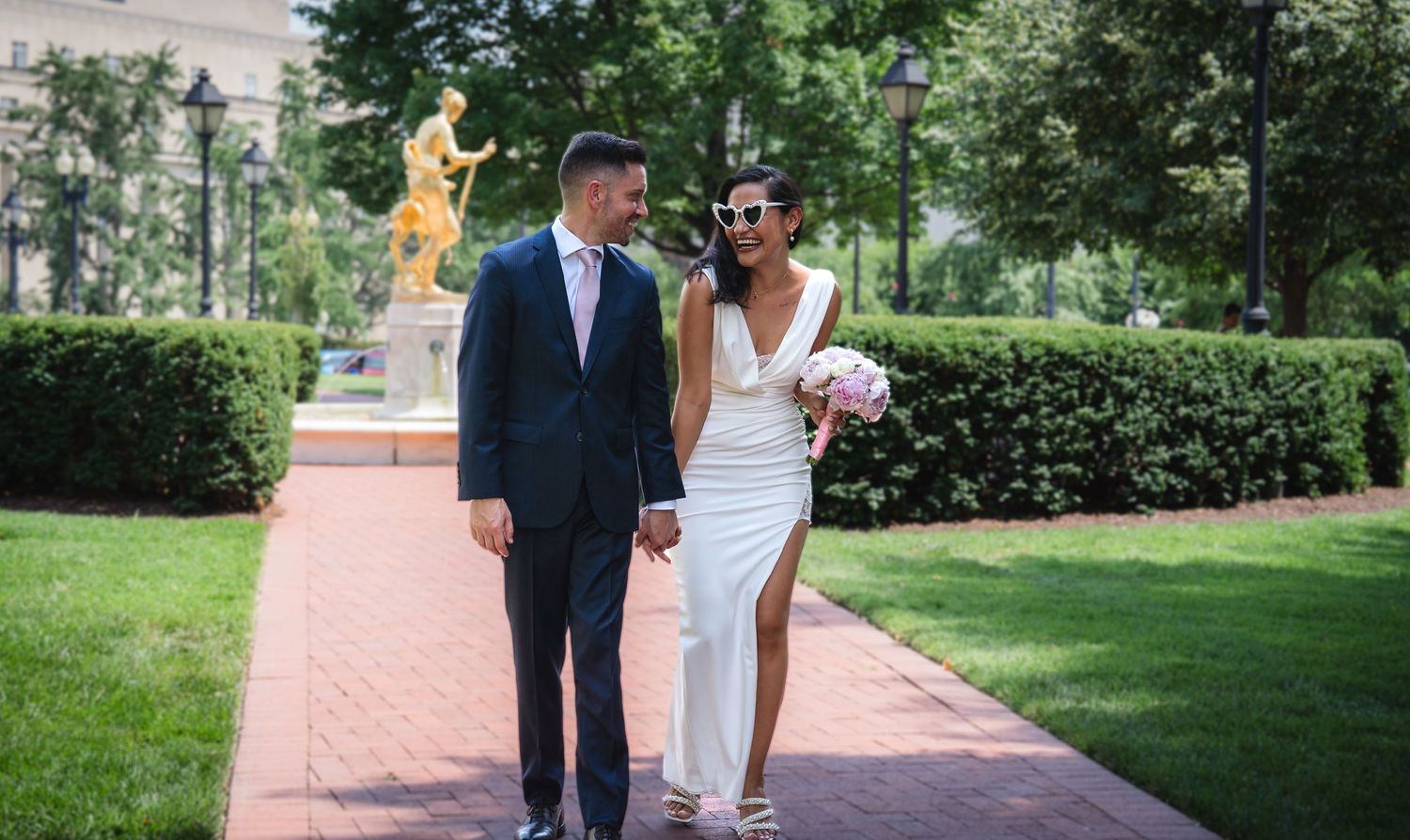 Elegant bride and groom walking hand-in-hand along a brick path in a lush garden with a golden statue backdrop.