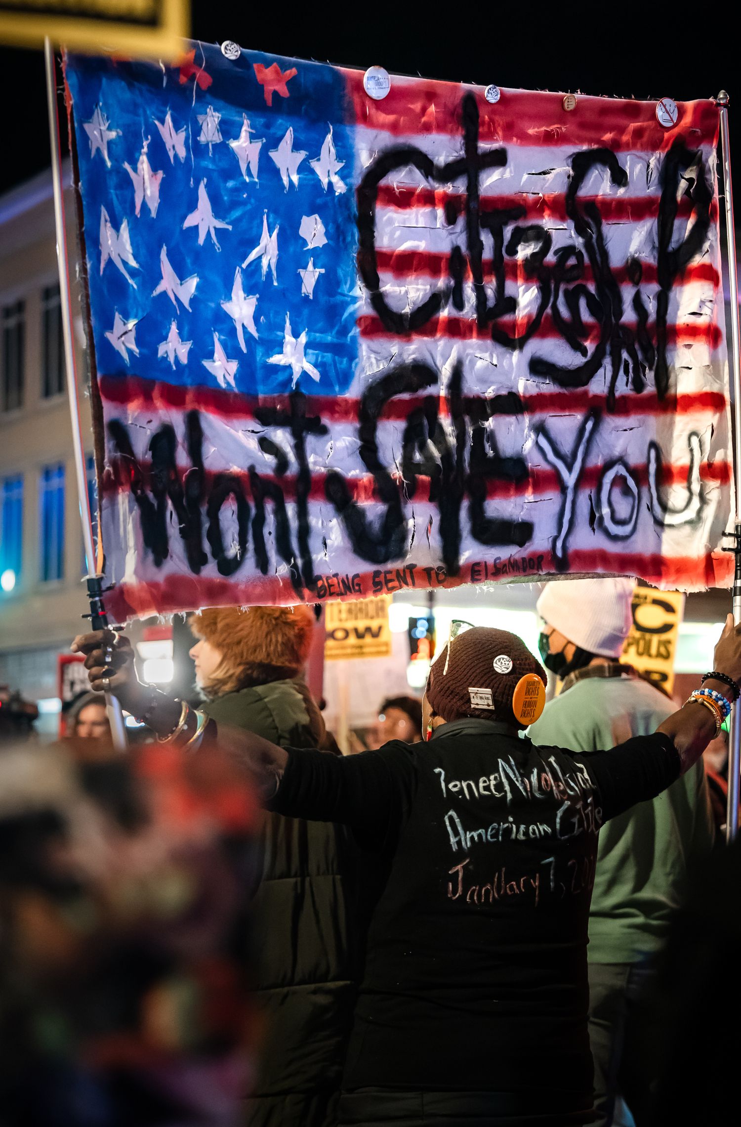 Protesters gather in front of a graffitied American flag banner during a nighttime demonstration.