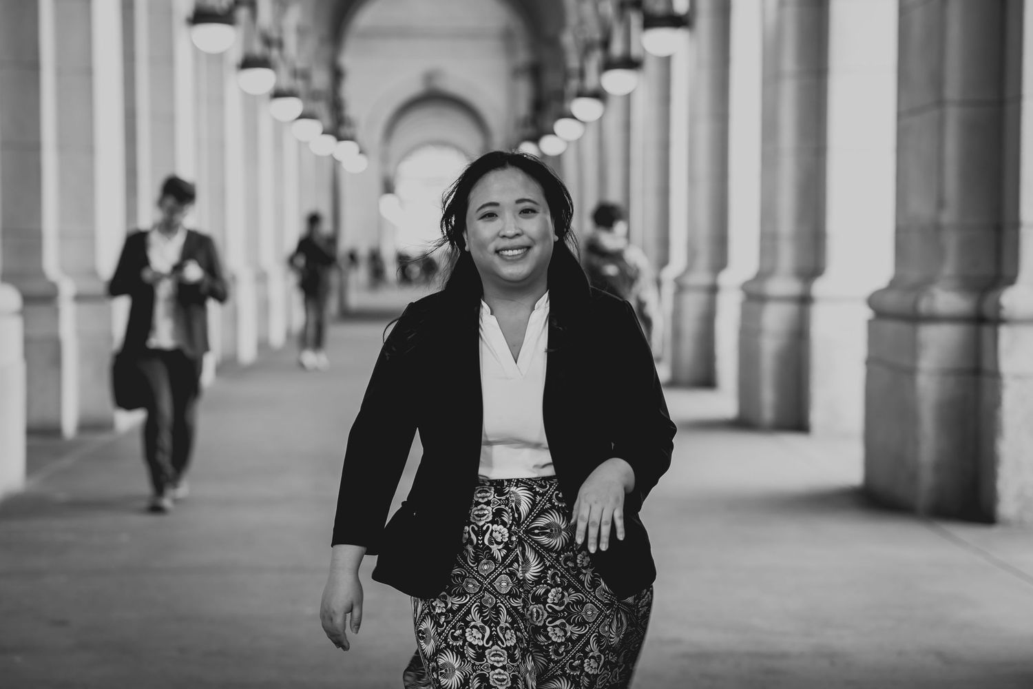 Black and white photo of a smiling woman in a blazer and patterned skirt walking through an elegant columned corridor.