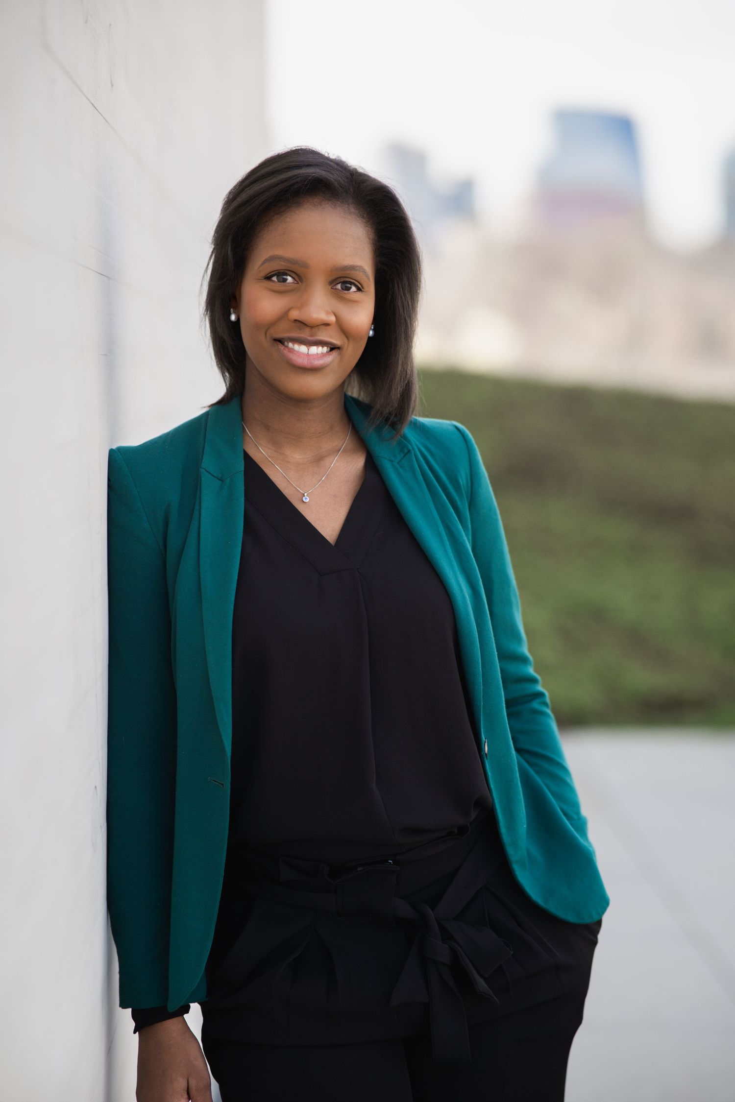 Person in teal jacket and black dress stands elegantly against white backdrop.