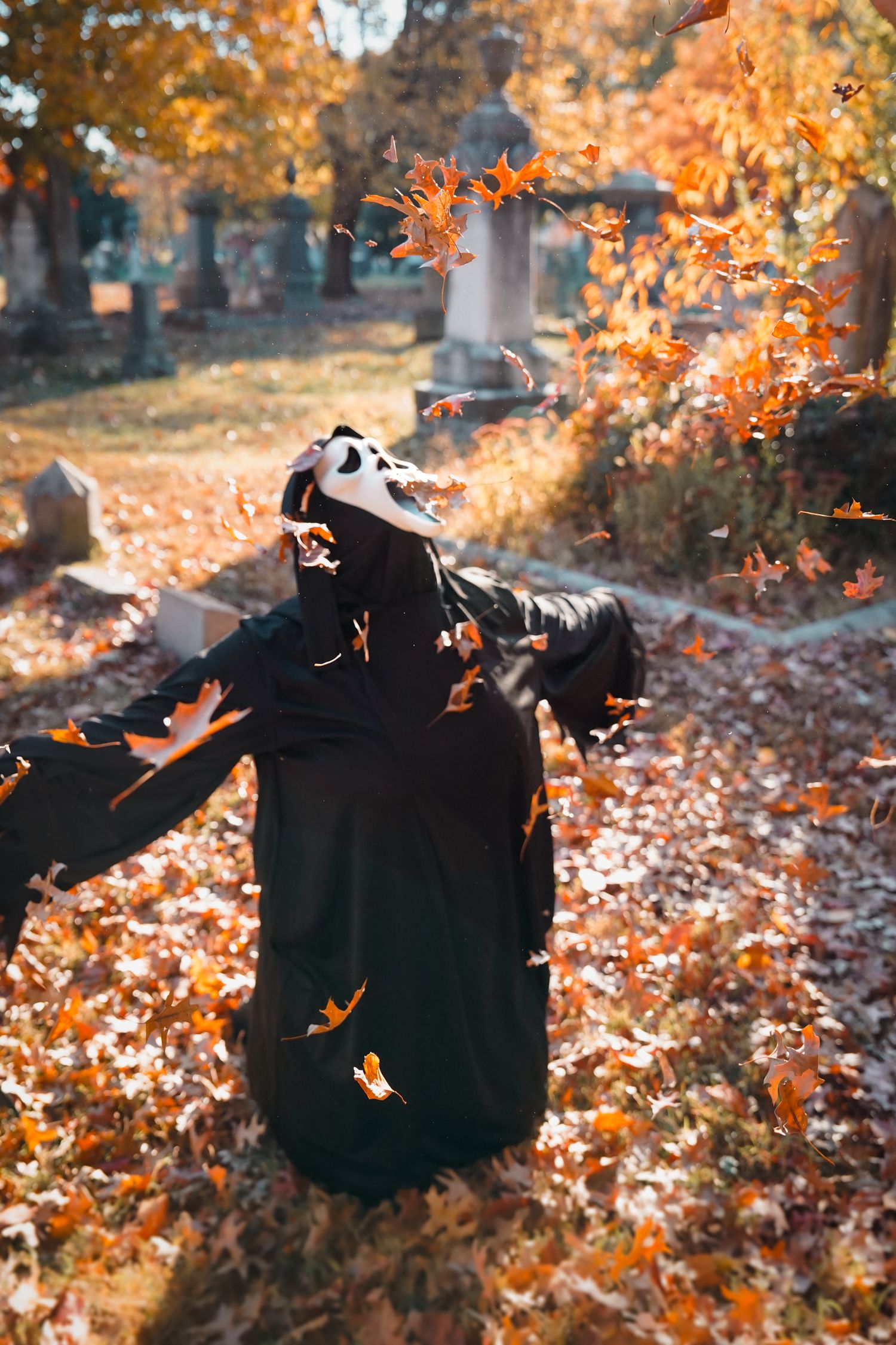 Figure in Ghostface mask spinning among falling autumn leaves in cemetery.