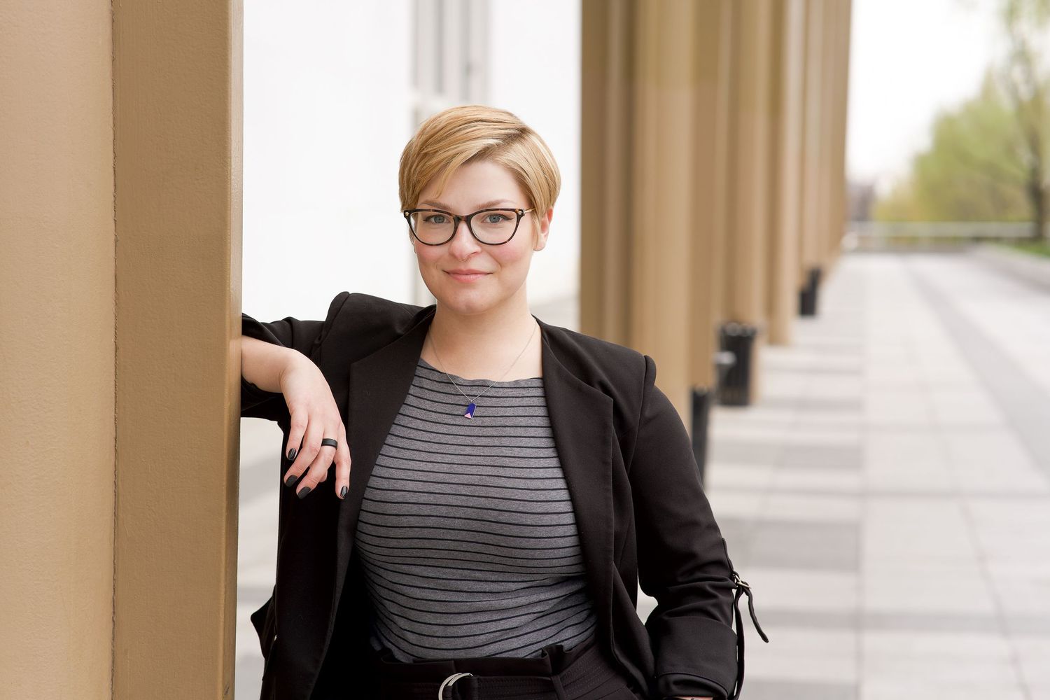 Professional portrait in business attire with striped top and black blazer leaning against a column outdoors.