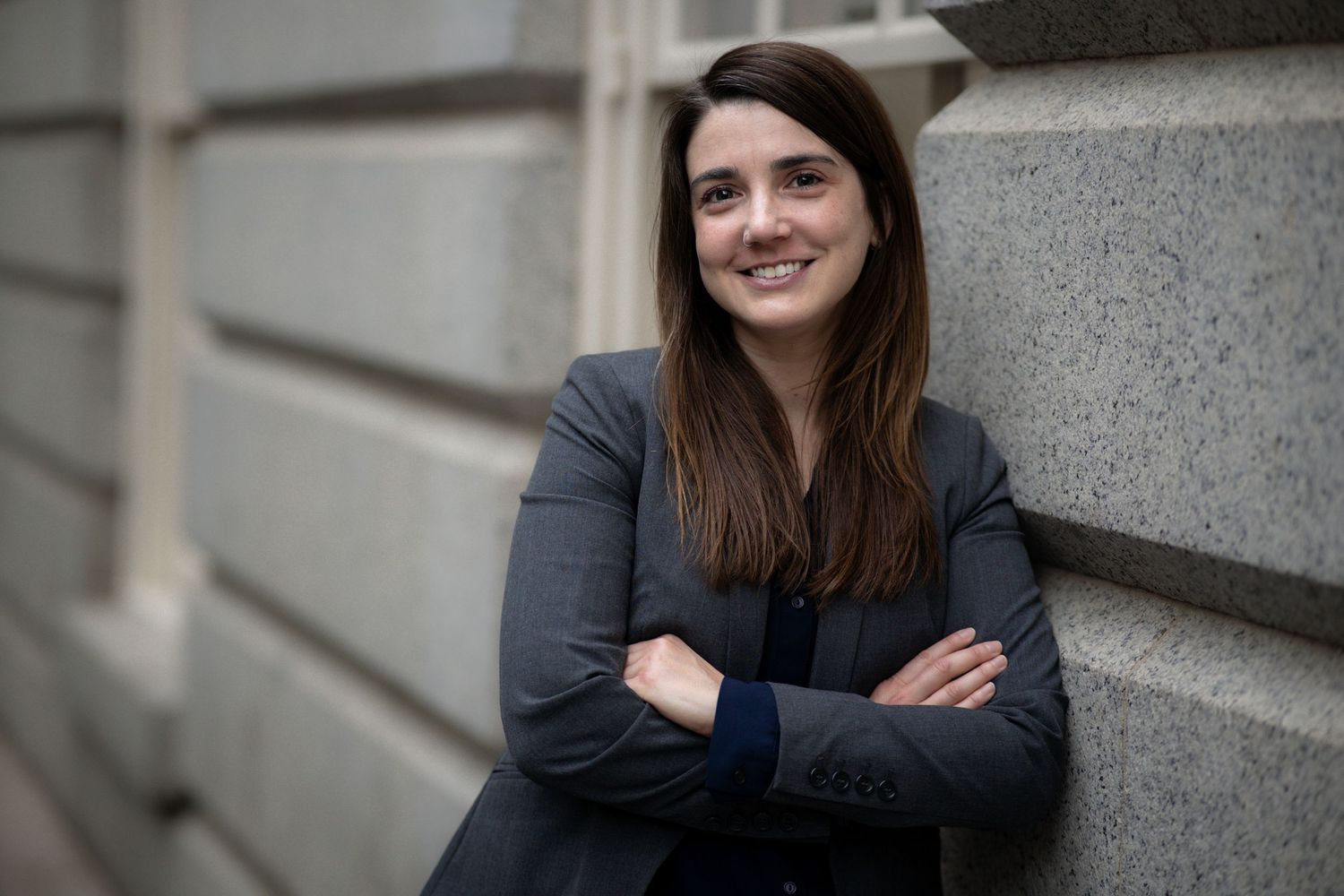Professional with long dark hair in grey blazer leaning against stone wall with arms crossed.
