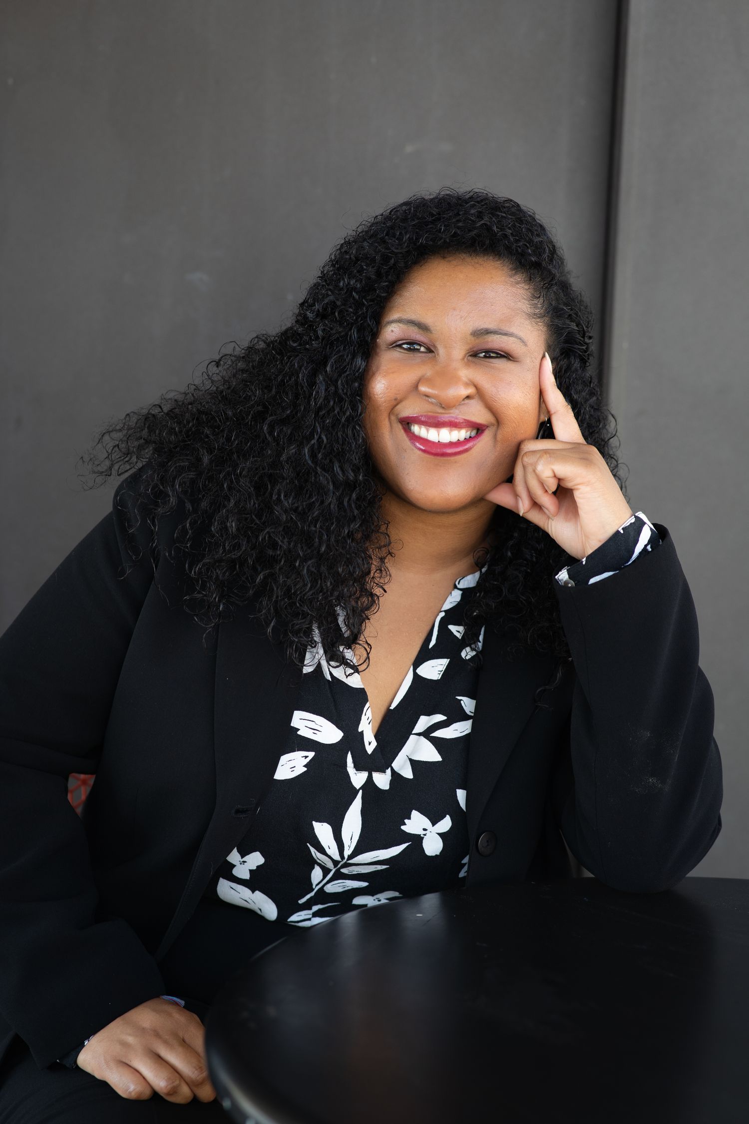 Smiling Black woman with curly hair wearing a black blazer and floral blouse, posing confidently in a professional portrait.