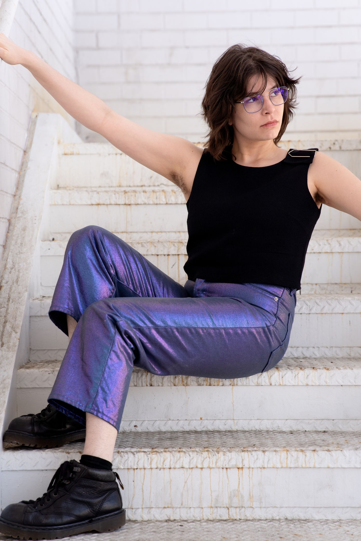 Person sitting on white stairs wearing shiny purple pants, black tank top, glasses, and black boots with one arm extended.