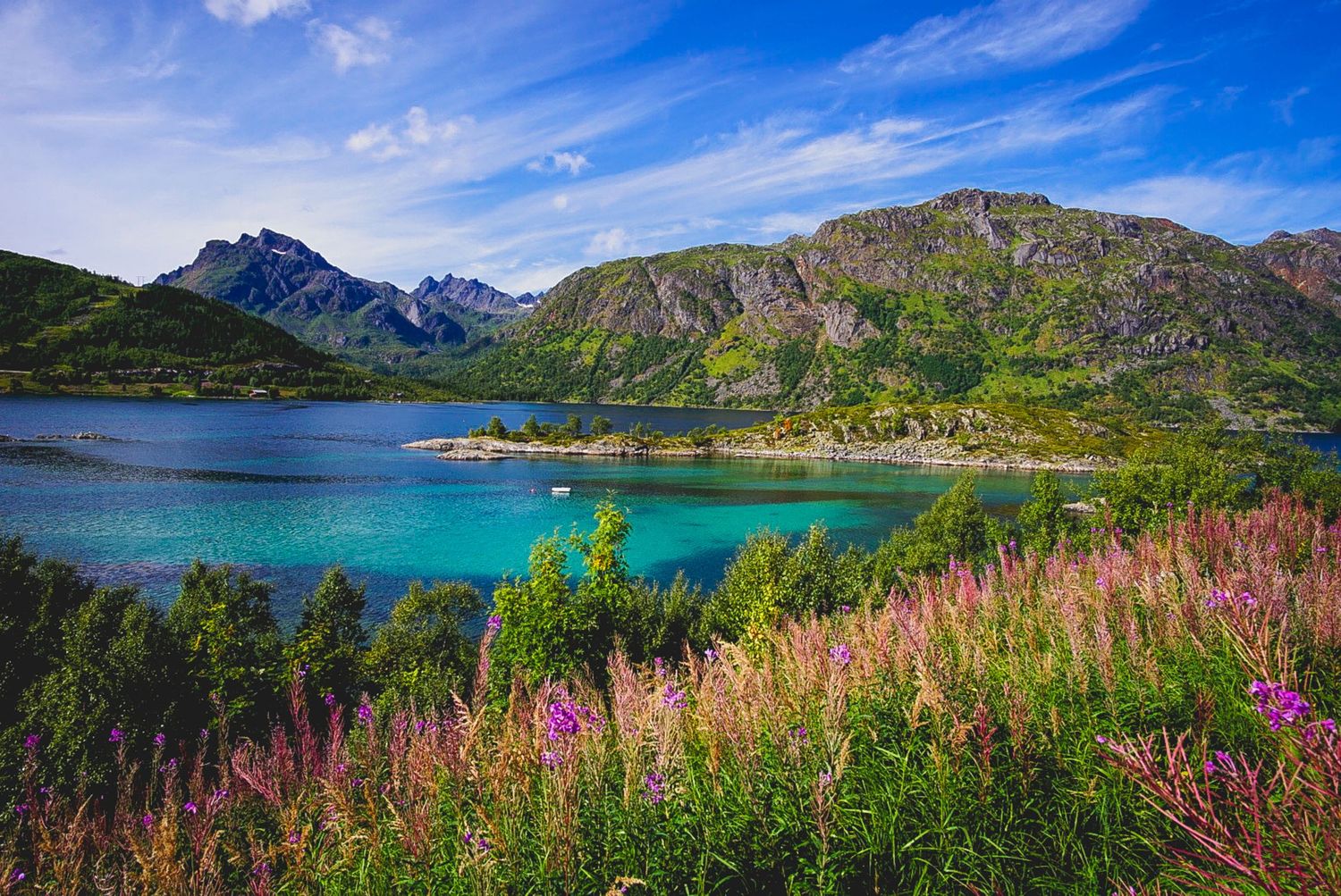 Turquoise mountain lake surrounded by peaks and blooming pink wildflowers in summer.