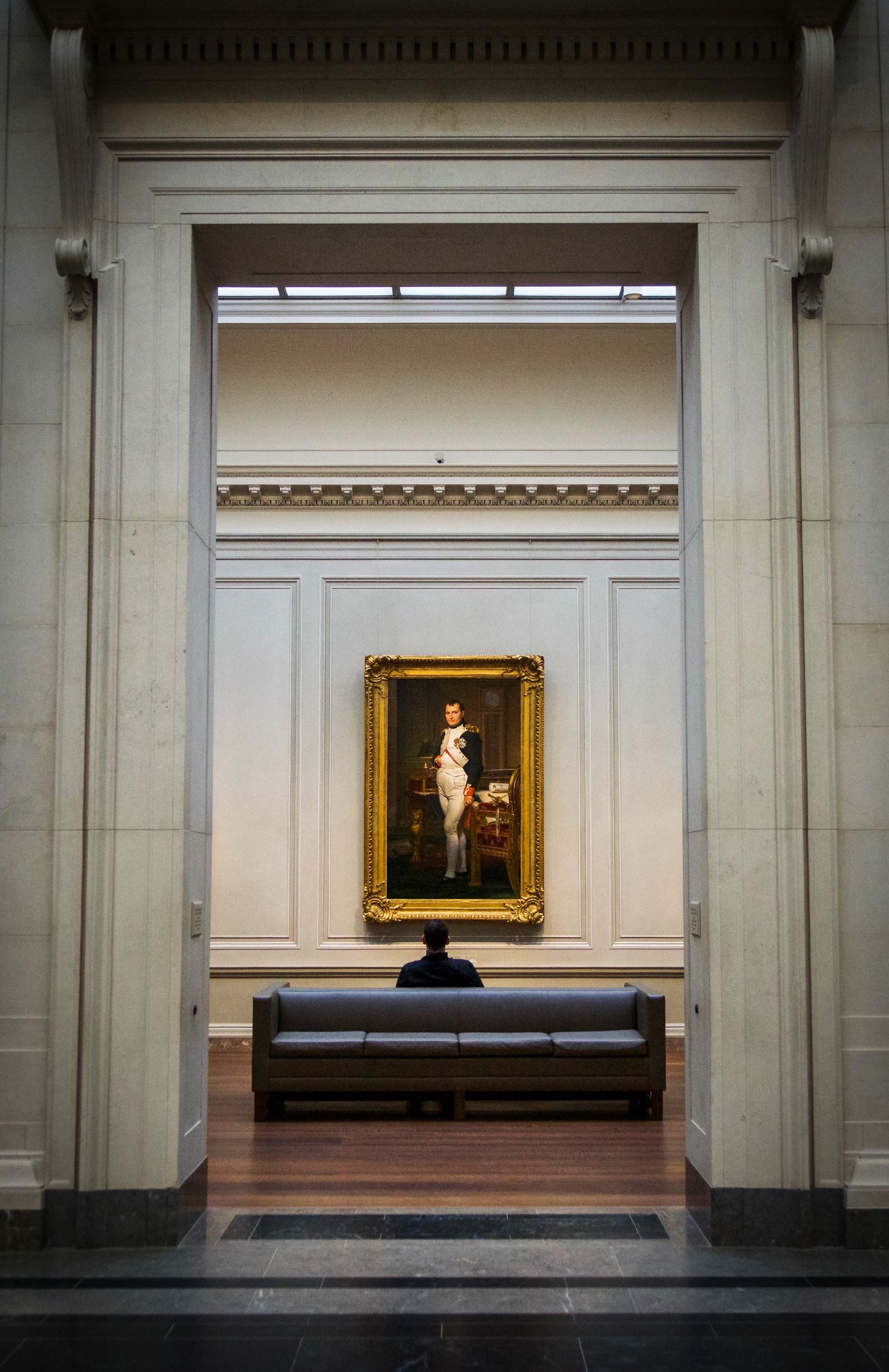 Museum gallery view showing a classical portrait painting in gold frame with bench below.
