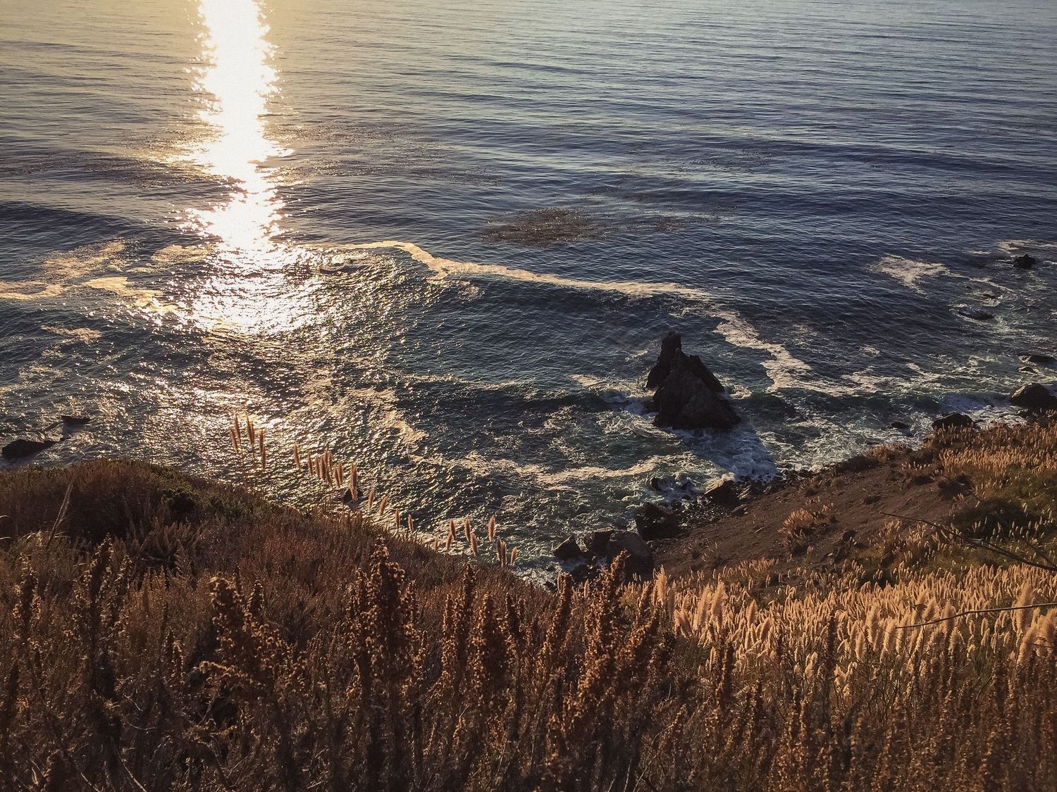 Golden sunlight reflecting on ocean waves with rugged coastal cliffs in the foreground.