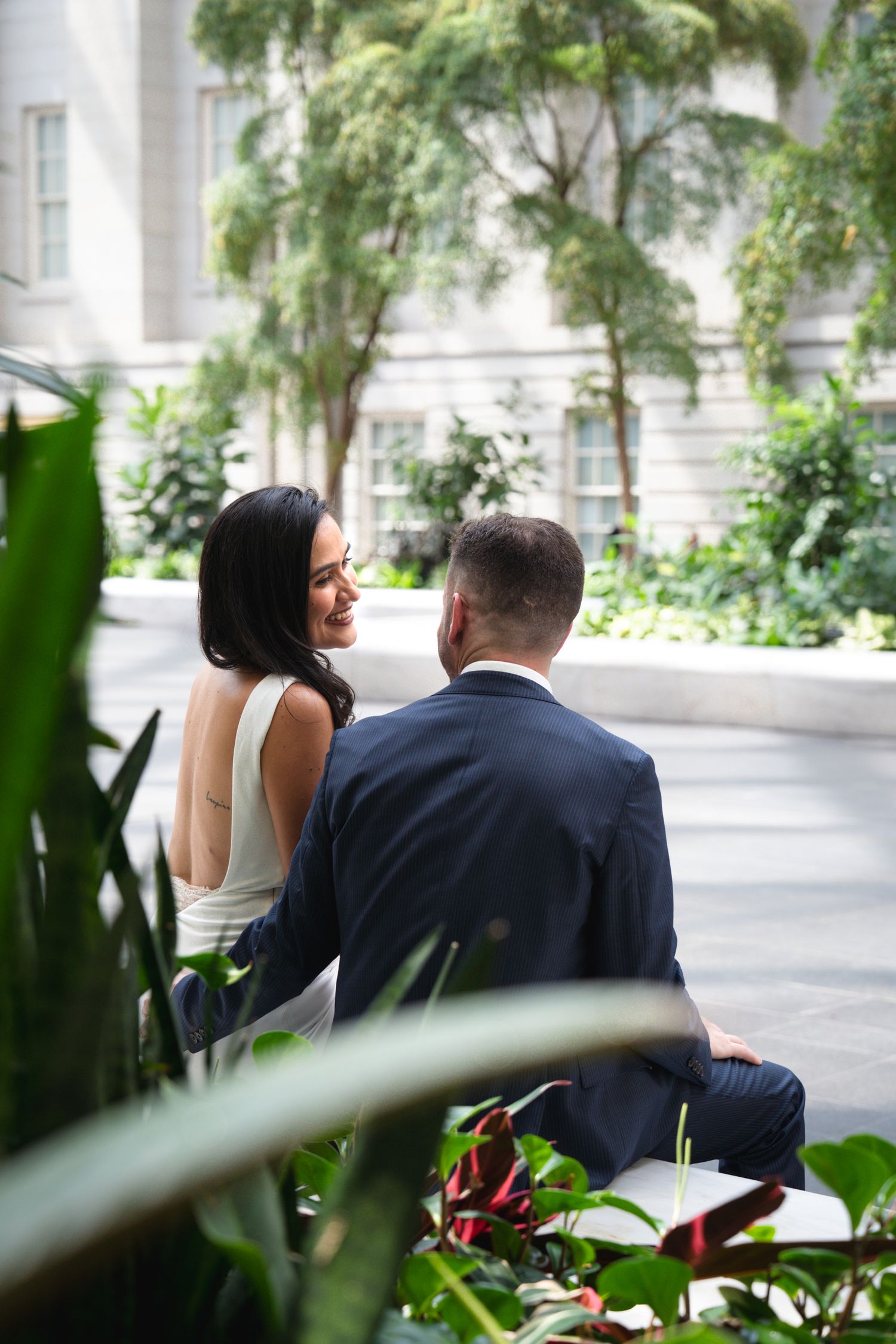 A bride in white dress smiles at groom in navy suit while seated among lush greenery in elegant courtyard.