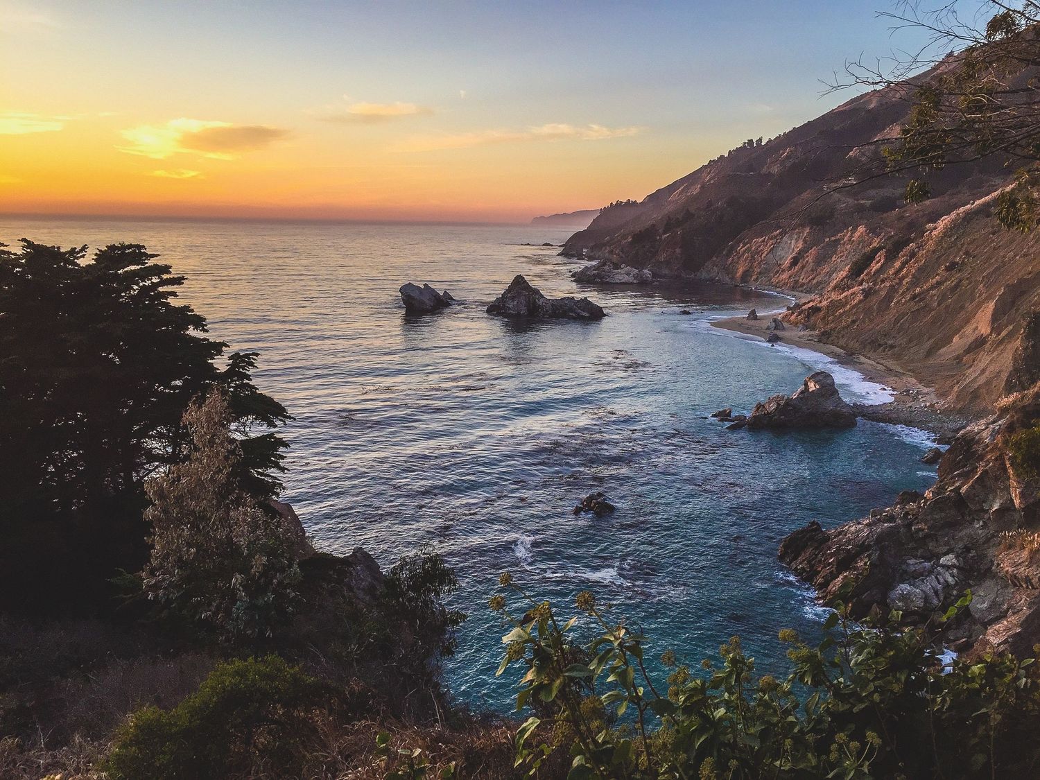 Dramatic sunset over Big Sur coastline with waves crashing against rocky cliffs.